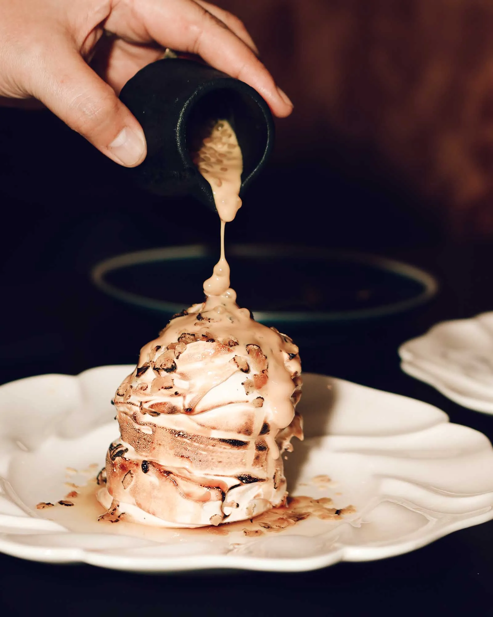 Hand is pouring caramel sauce over a chocolate-chip cupcake on a white plate, close-up view.