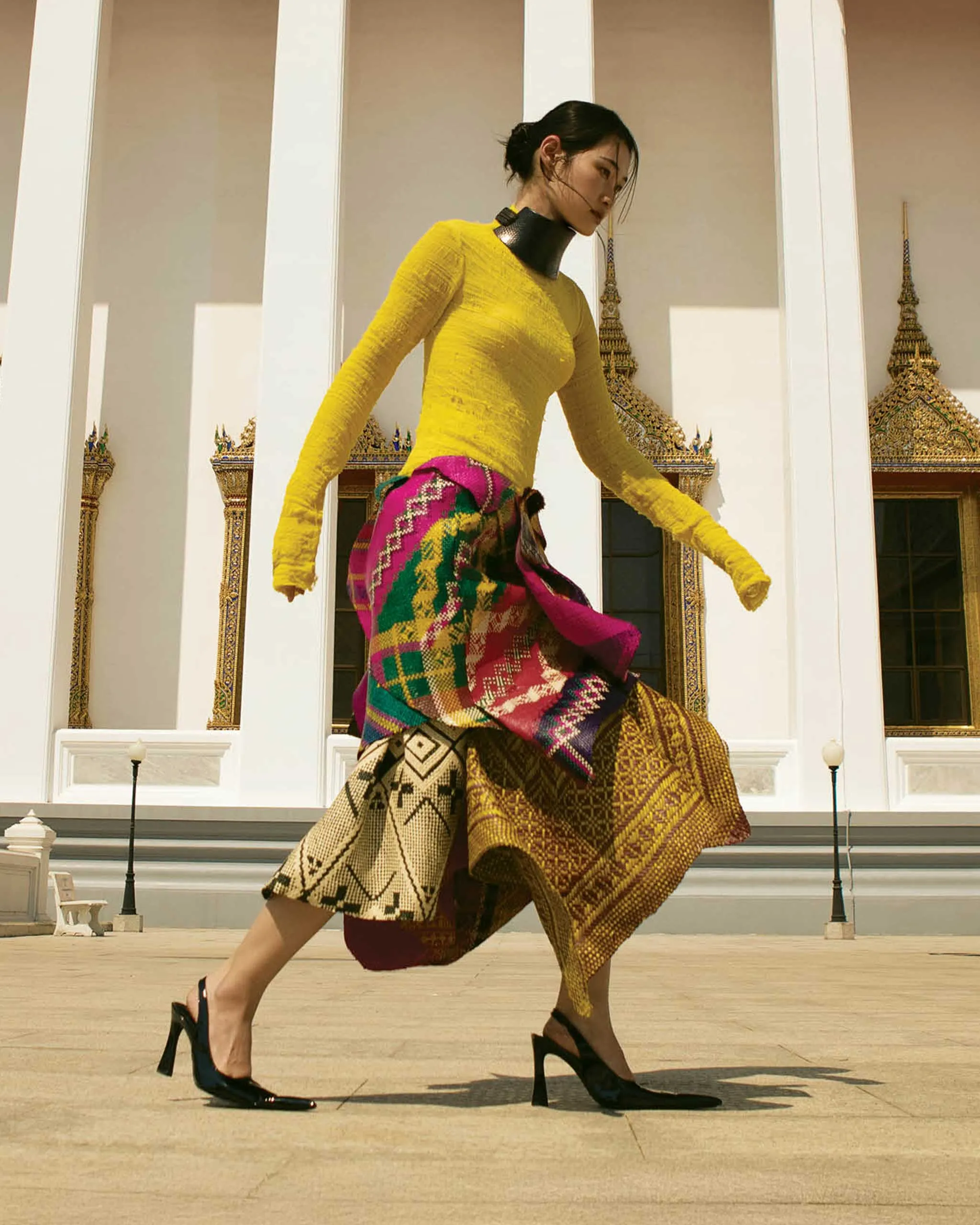 Woman in a bright yellow long-sleeve top and a colorful patterned wrap skirt walking past a temple with white columns and gold decorations.