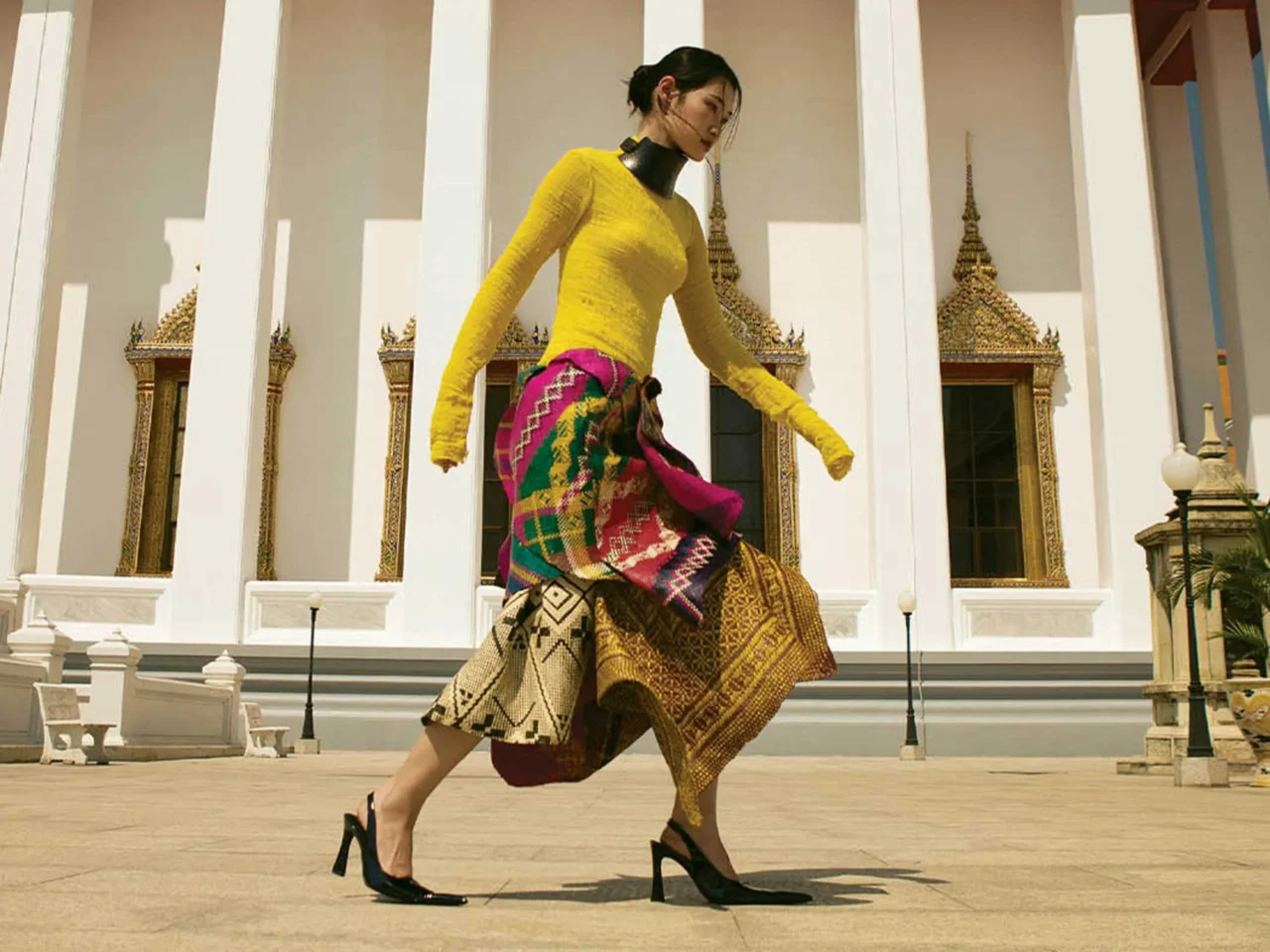 Woman in a bright yellow top and multicolored patterned skirt walking past a white temple with gold ornate windows and columns in the background.