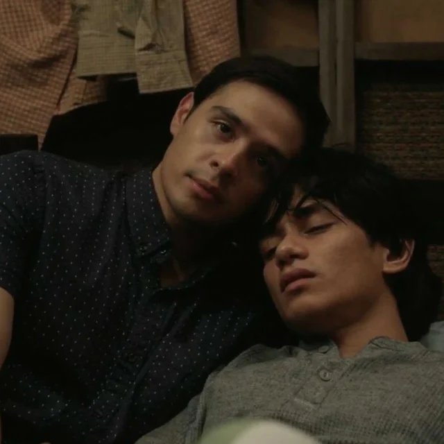 Two men lying close together on a couch, with one resting his head on the other's shoulder.