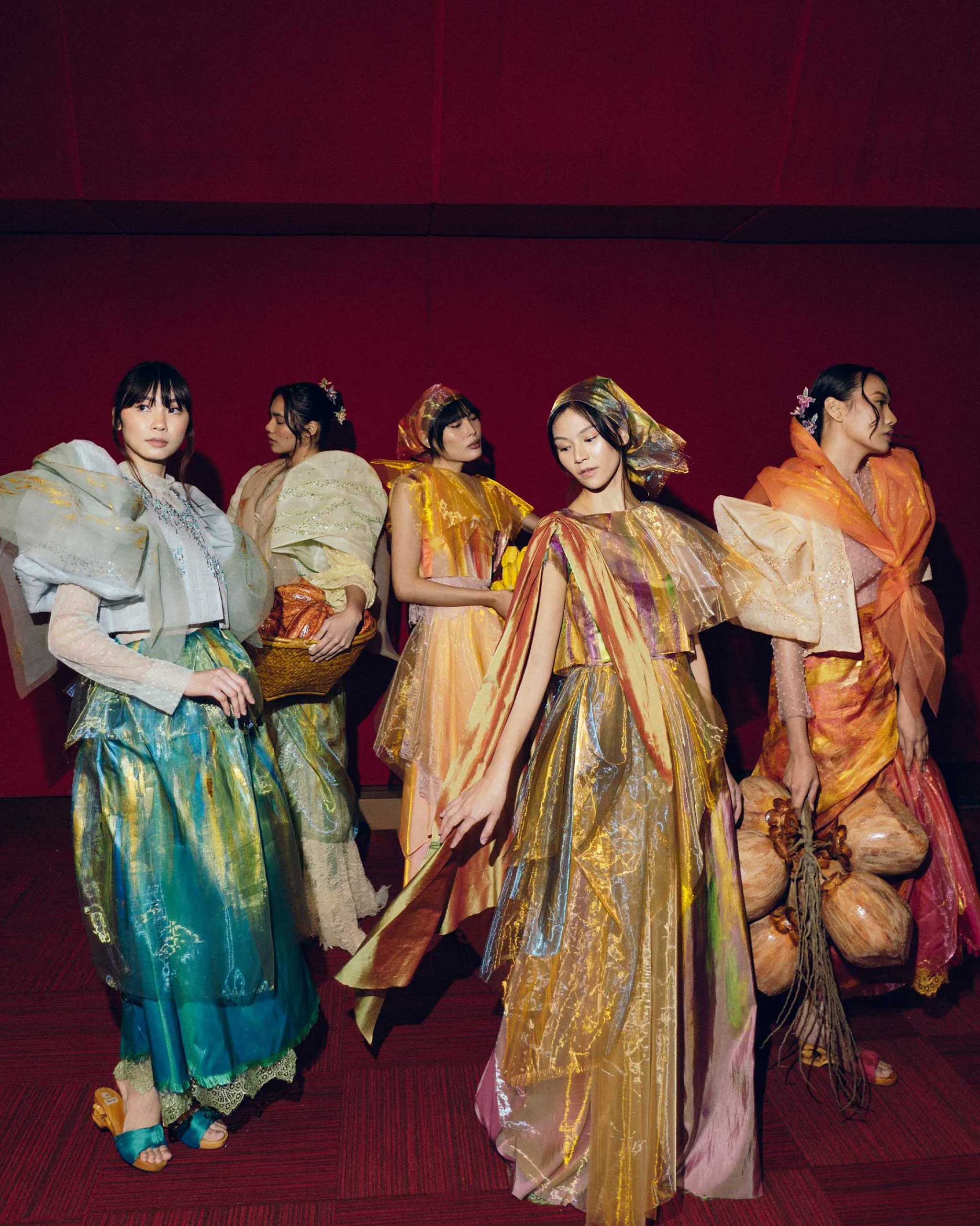 Models wearing vibrant, flowing gowns pose together against a deep red backdrop. Five women in iridescent dresses create a colorful fashion-show scene.