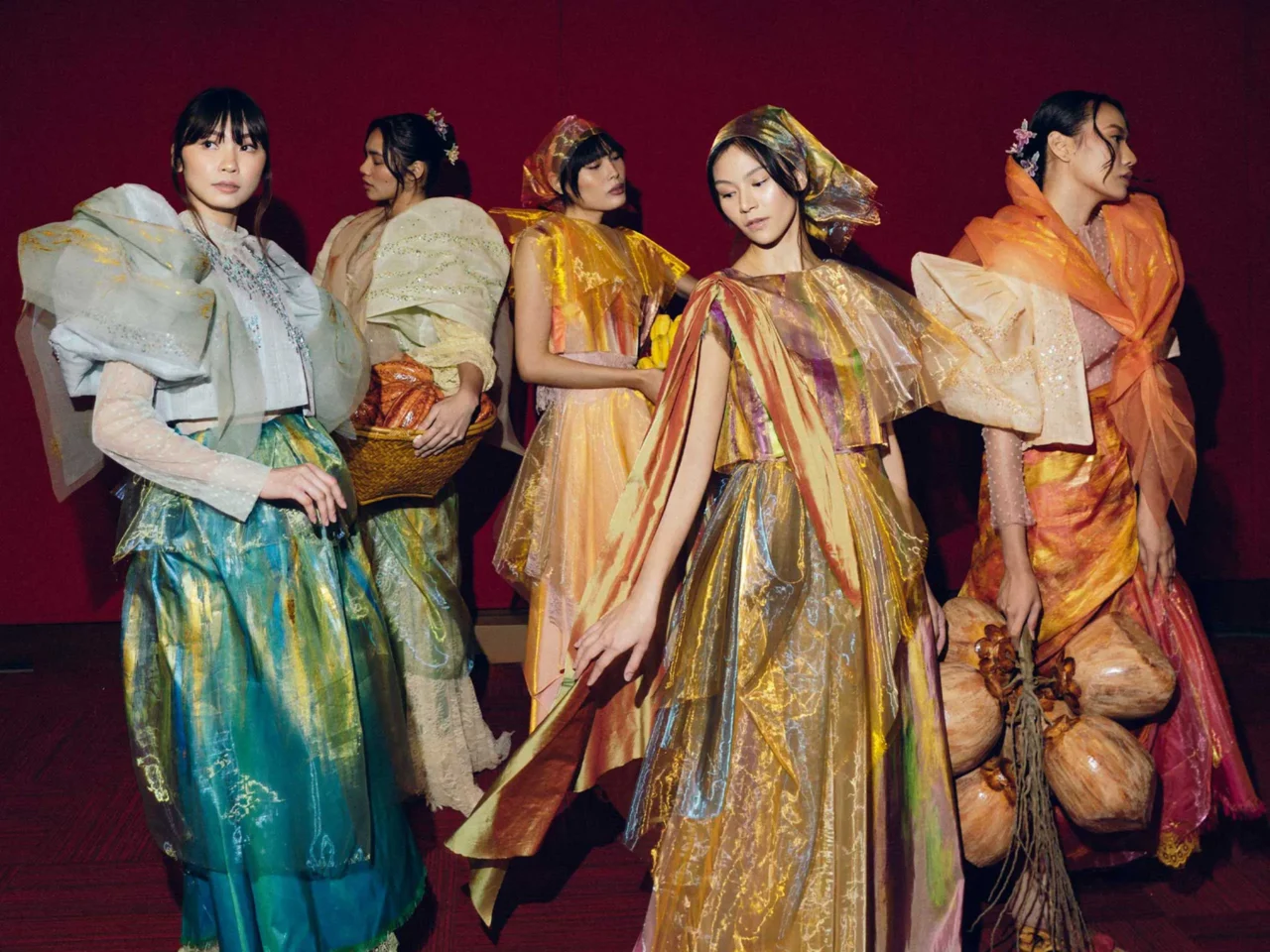 Group of models in colorful, flowing gowns on a stage with a red curtain behind them, posing as a fashion show walk.
