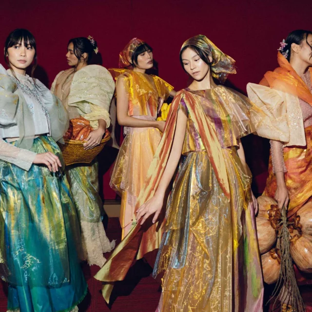 Five women in colorful, flowing gowns pose together on a stage with a dark red curtain behind them.