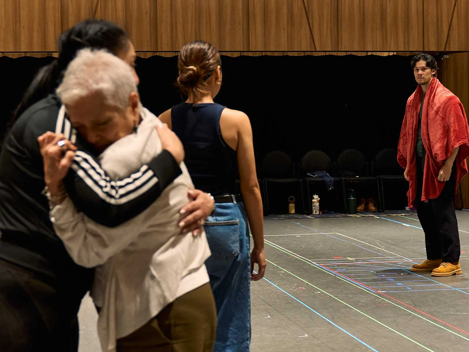 Two people embrace in the foreground as a man wrapped in a red towel waits on a stage in the background backstage area.