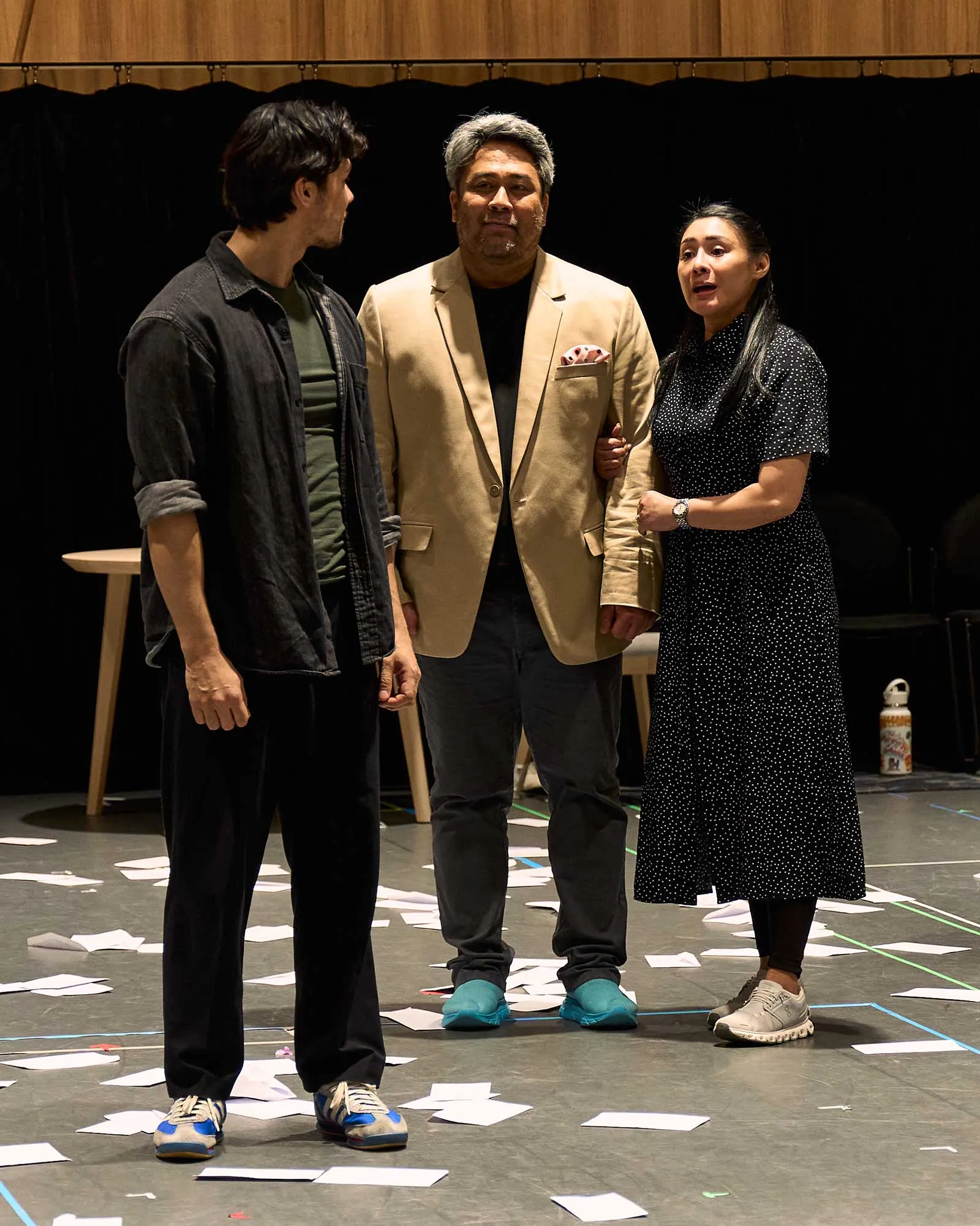 Three adults stand on a stage with scattered papers on the floor; center man wears a tan blazer, flanked by two others.