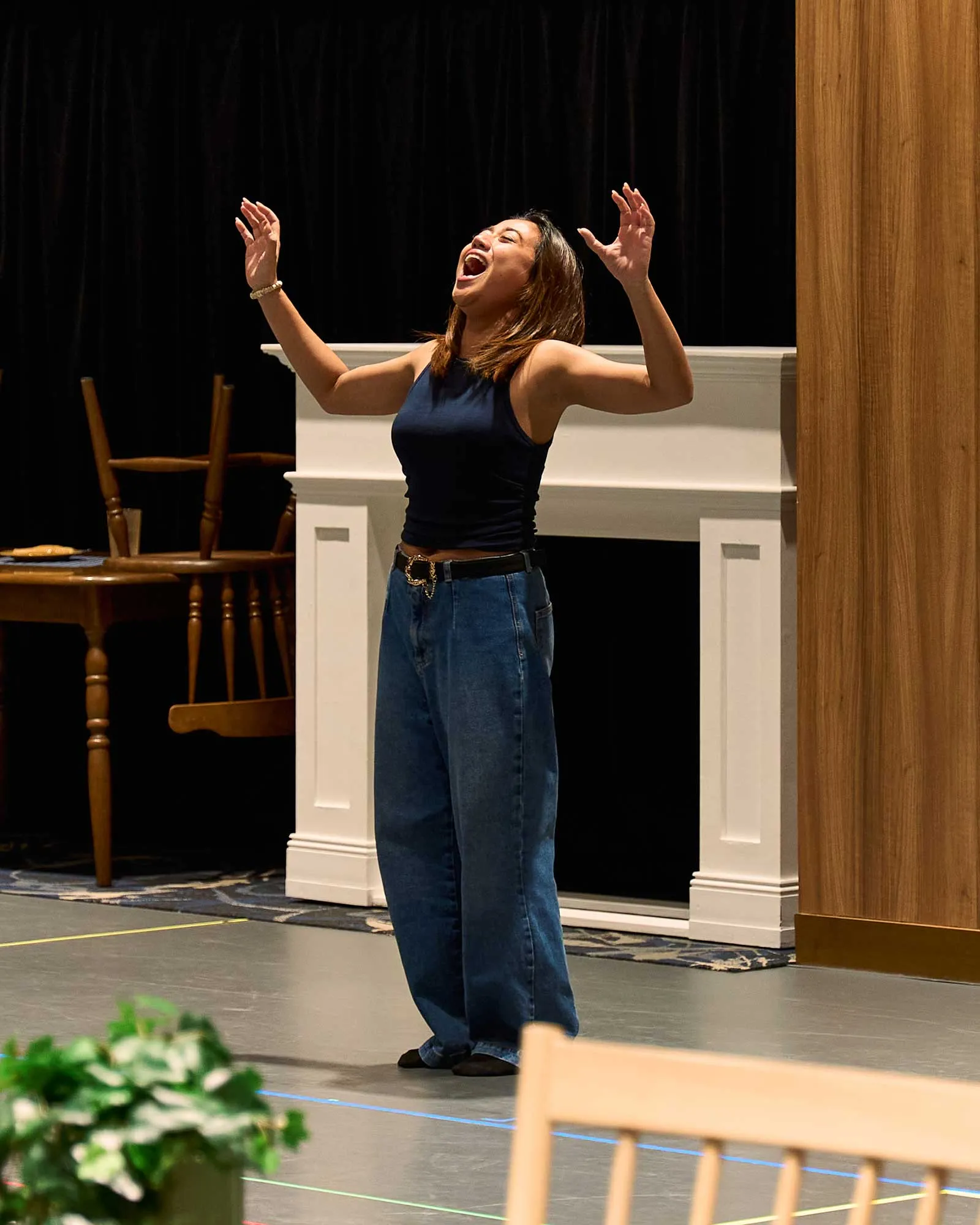 Woman on a stage singing with arms raised, wearing a navy sleeveless top and jeans, in front of a white piano and dark backdrop.