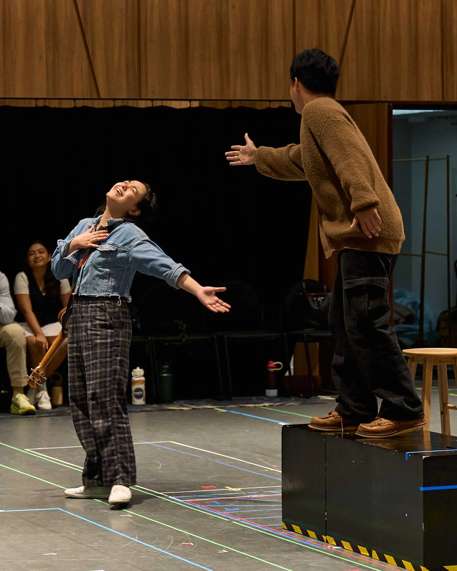 Two performers on a stage reach toward each other while a smiling woman in denim watches from the left audience.