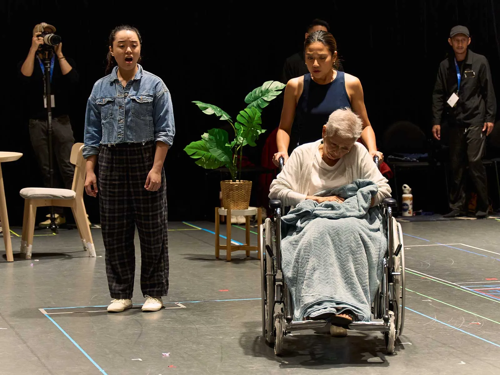 Stage scene with a young singer in a denim jacket standing beside an elderly woman in a wheelchair being guided by a helper; backstage crew and a potted plant visible in the background.