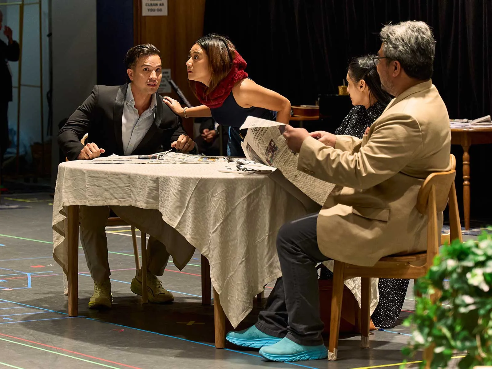 Four people sit at a table covered with a tablecloth, reviewing papers; a woman in a red scarf leans toward a man in a blazer while others read a newspaper.