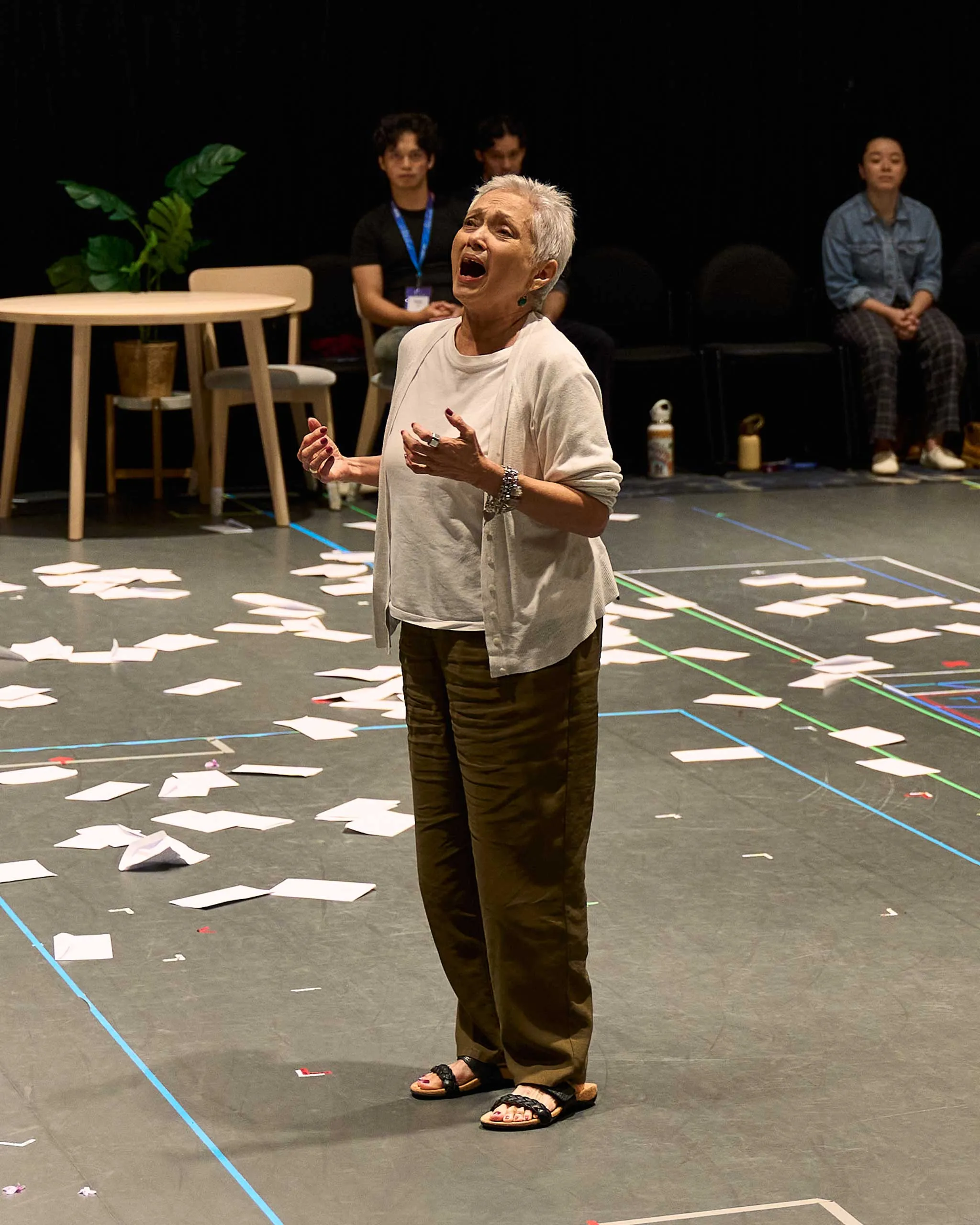 Older woman with short gray hair singing passionately in a rehearsal space, papers scattered on the floor and taped lines on the floor marking areas behind her