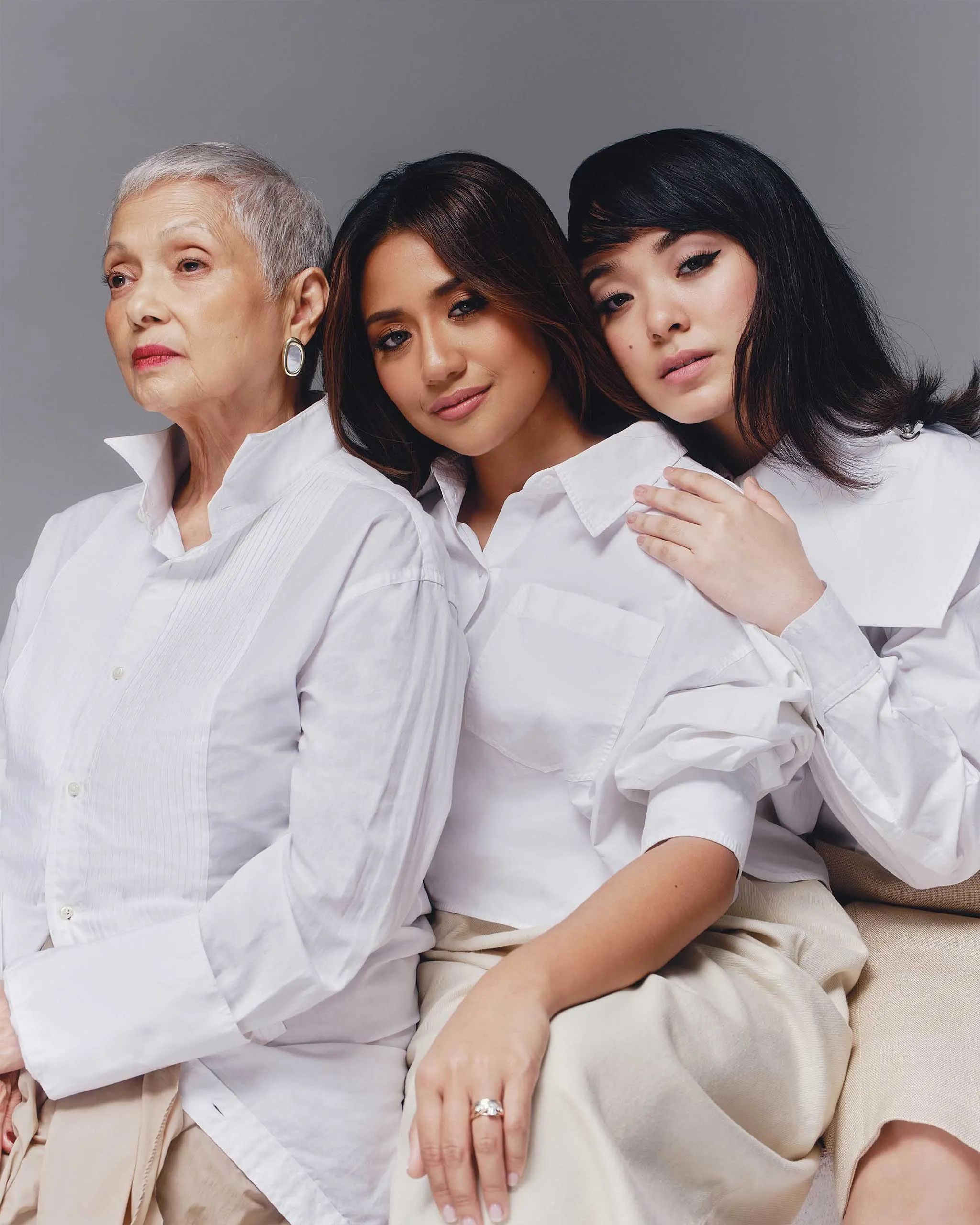 Three women of different ages in white shirts posing closely together against a gray backdrop.