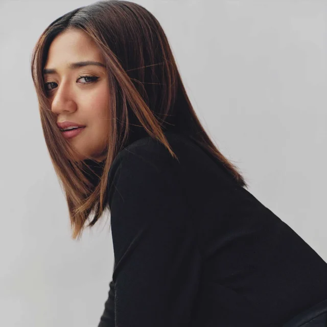 Close-up of a woman with long brown hair looking over her shoulder against a gray background, wearing a black top
