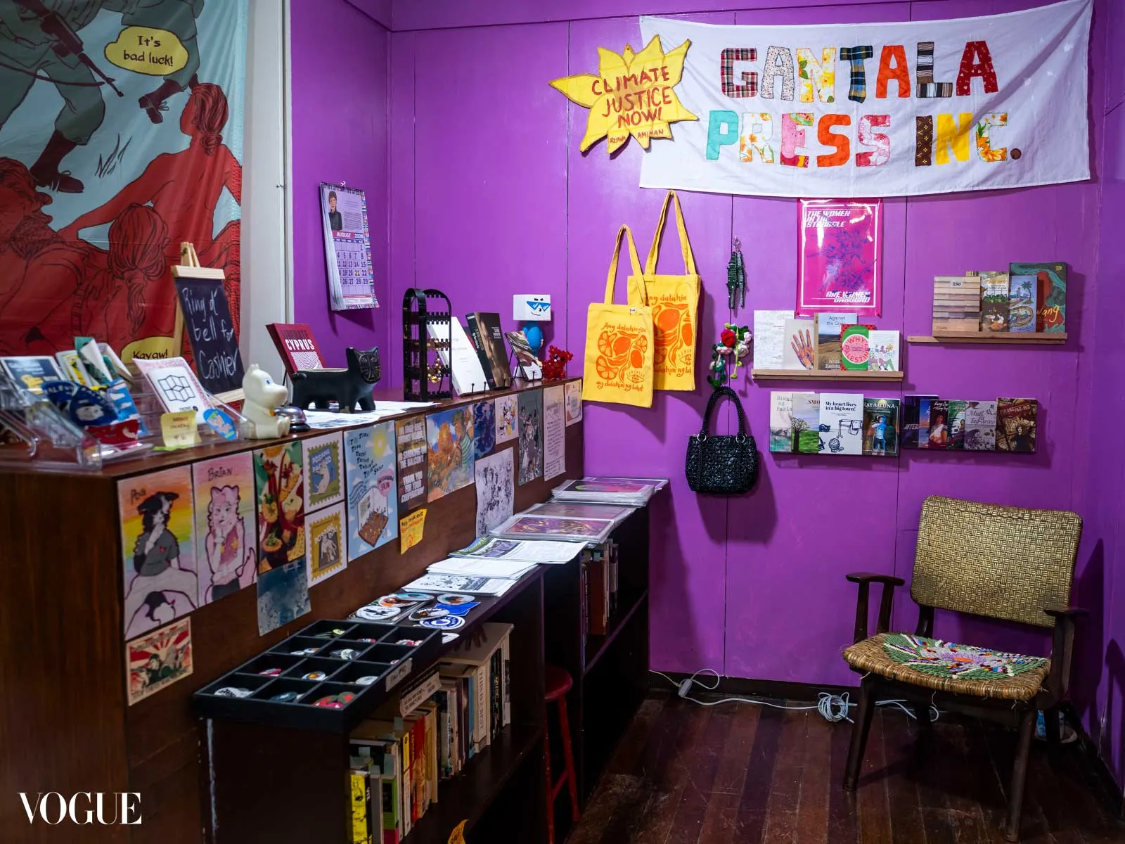 Vibrant bookstore nook with purple walls, shelves of books, postcards, and colorful tote bags hanging; a woven chair sits nearby under a large banner that reads 'Gantala Press In'.