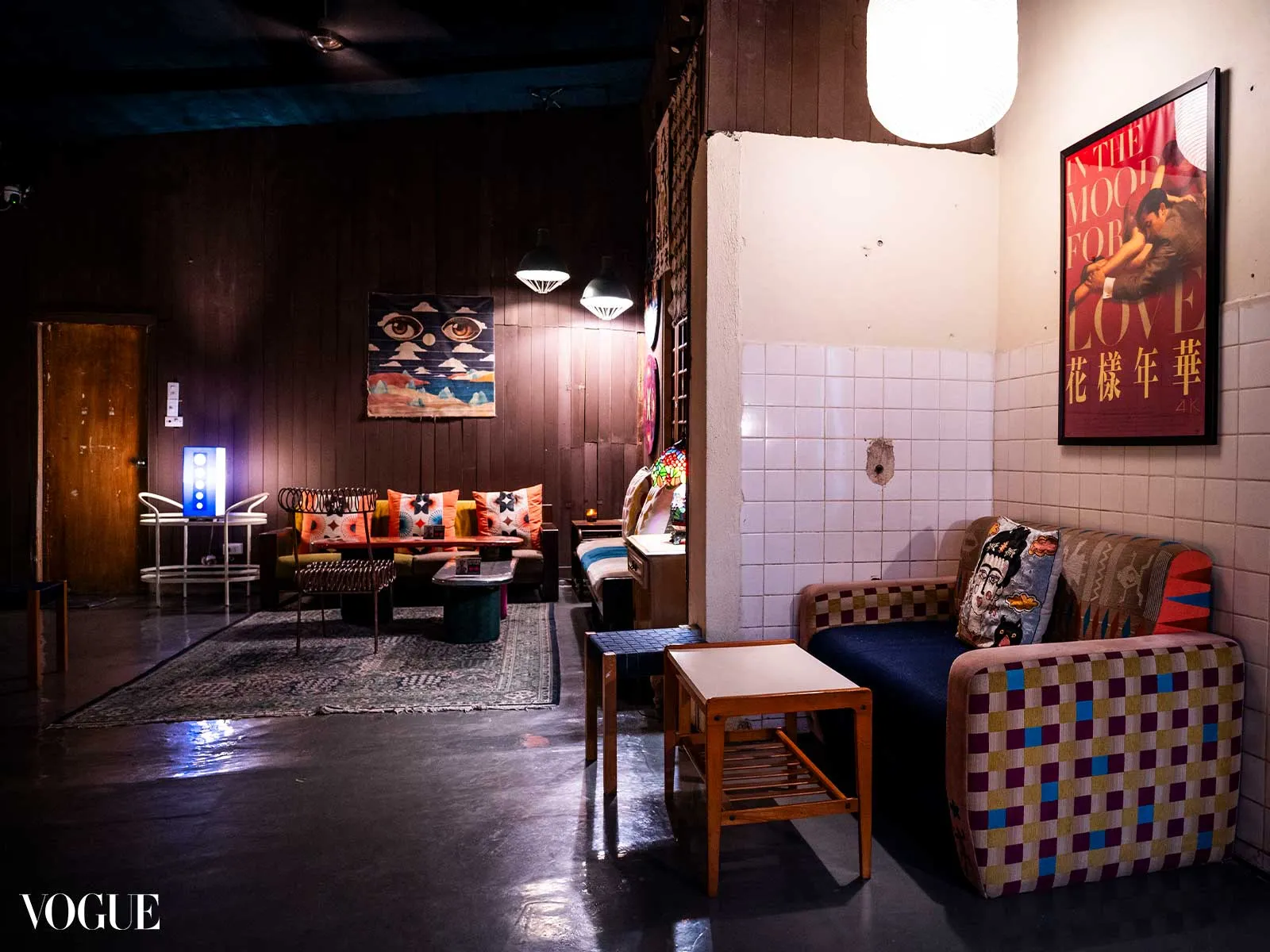 Dimly lit retro lounge with colorful patchwork couches, wooden walls, and framed art; Vogue watermark in the corner.