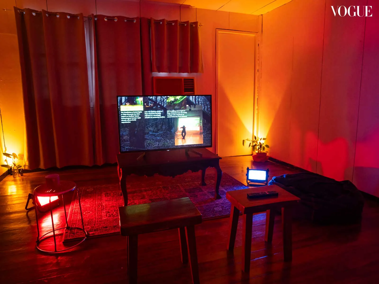 Dimly lit living room with red-orange and blue ambient lighting, TV on a stand showing a dancer, framed by curtains and a rug.