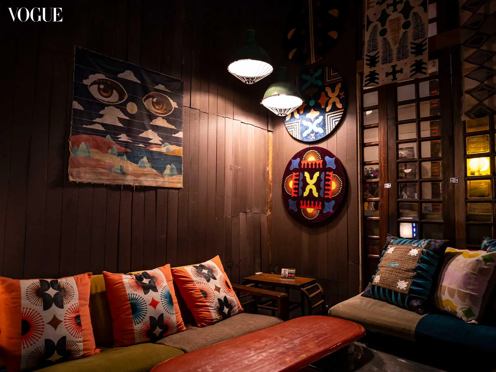 Cozy lounge with dark wooden walls, colorful patterned cushions, and round wall art; Vogue logo in the top-left corner.