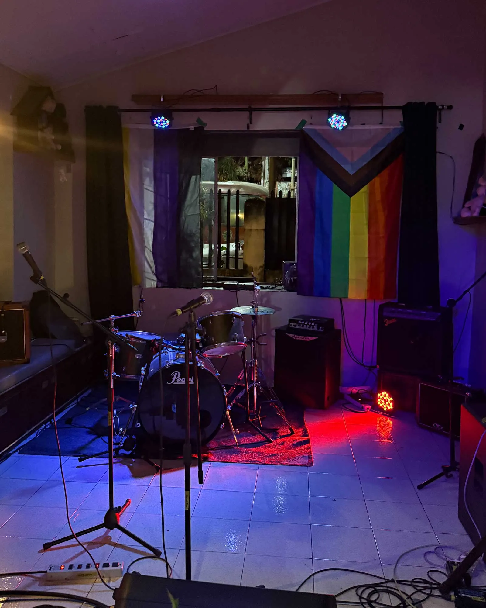 A small home stage with a Pearl drum set, microphones, and cables, lit by blue and purple stage lights, with a rainbow pride flag hanging on the right side.