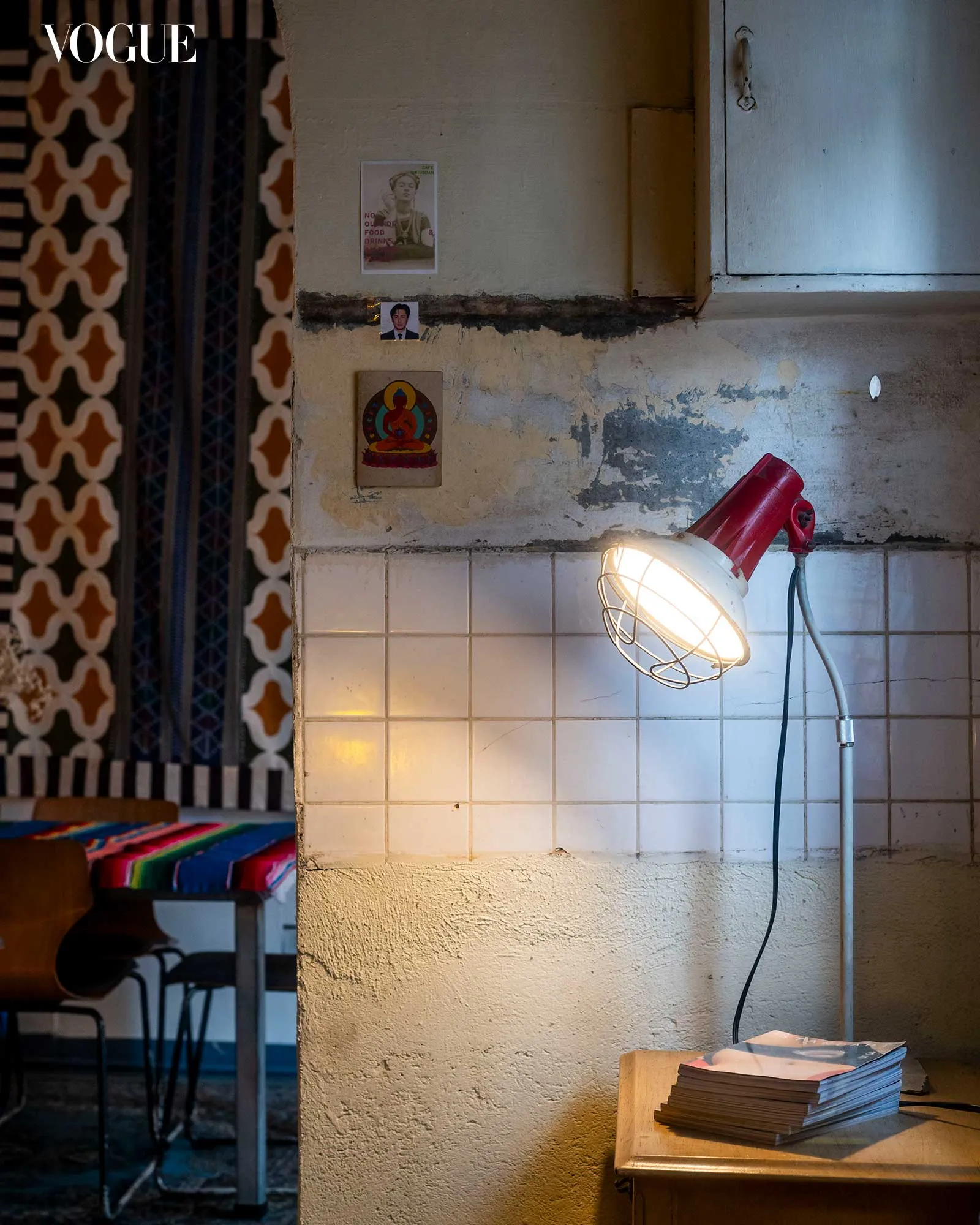 Vogue logo over a rustic interior: tiled wall with posters, a bright work lamp, and a stack of papers on a wooden table with a patterned backdrop on the left