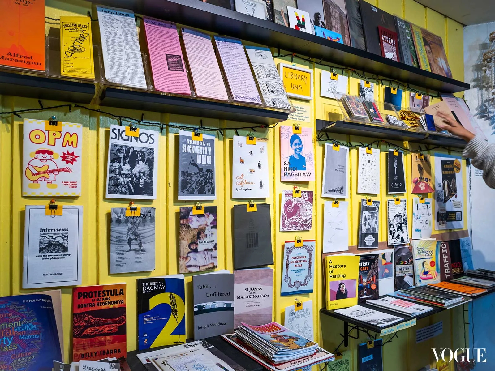 Colorful zine and poster wall on a bright yellow backdrop, with shelves above; a person’s hand reaches for a item on the right edge.