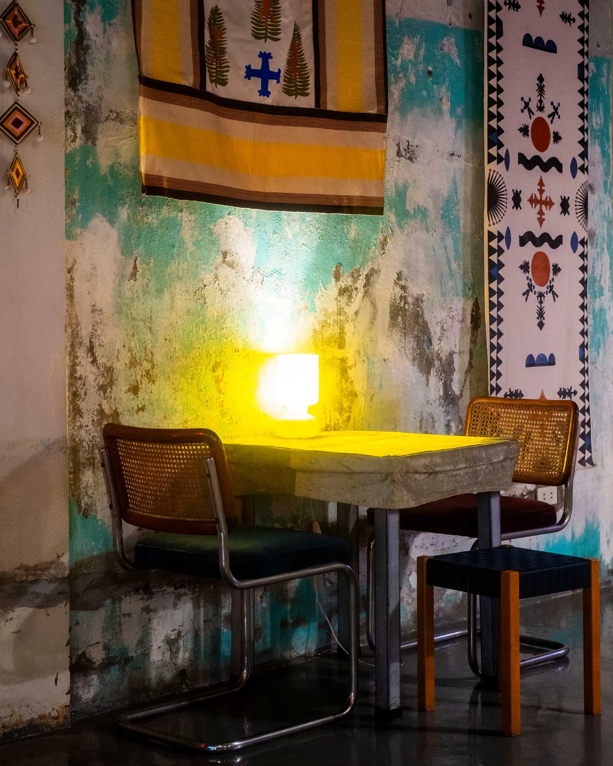 Cozy dining nook with a concrete table, woven chairs, and warm yellow lamp against a weathered turquoise wall with hanging textiles