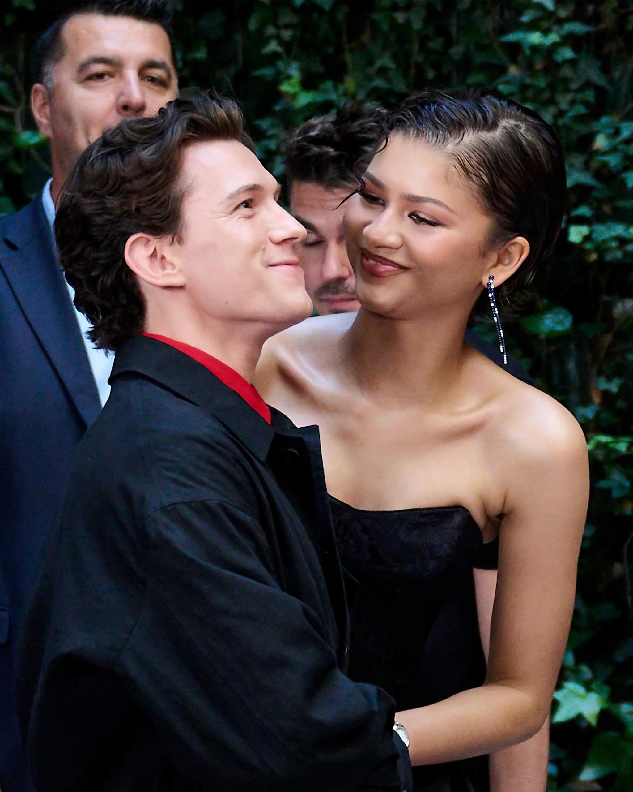 Couple in formal wear posing together outdoors, smiling, with green foliage in the background.