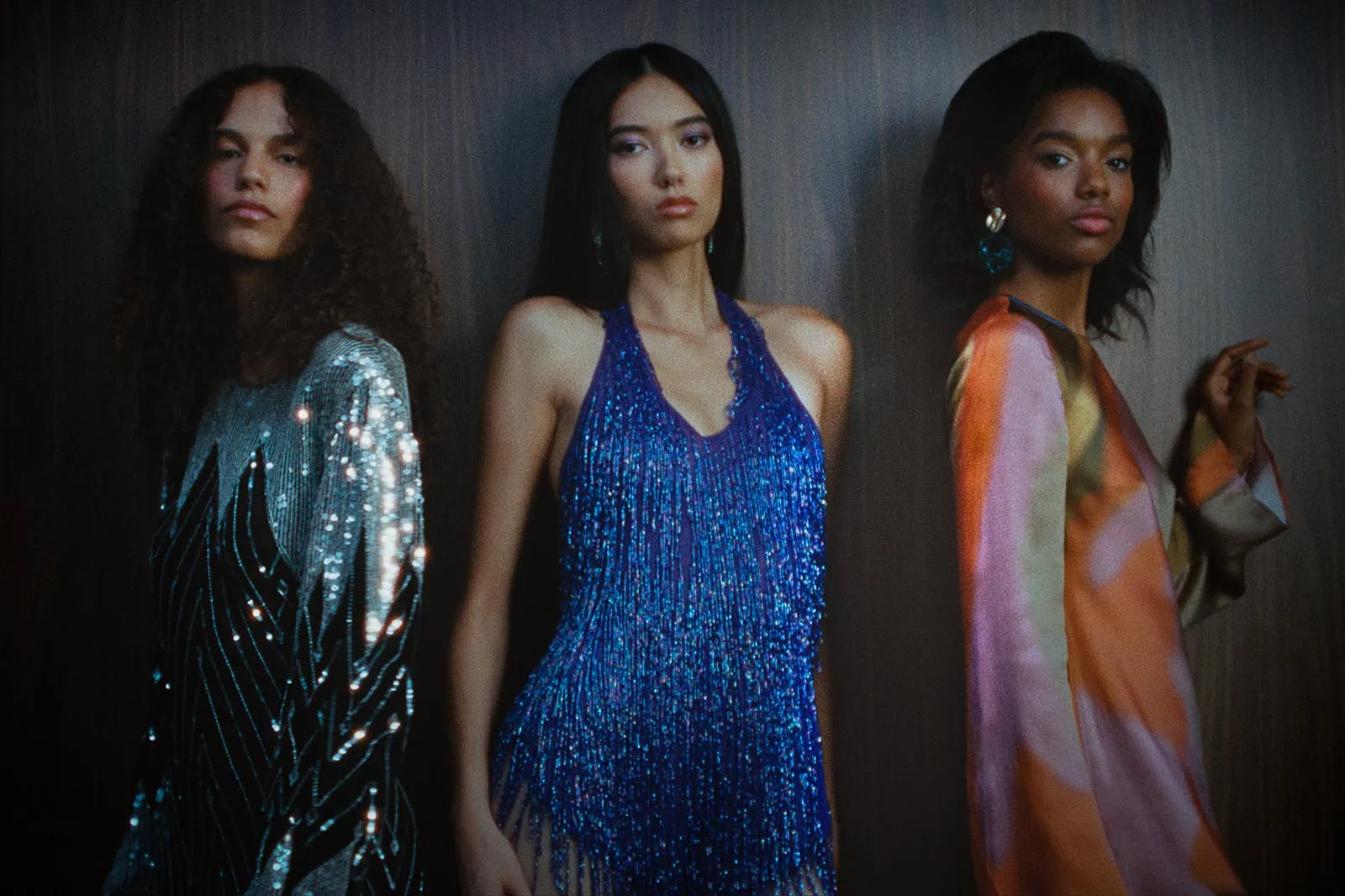 Three models stand side by side in glamorous outfits: left in a silver sequined dress, center in a blue fringed gown, right in a multicolored satin dress against a dark backdrop.