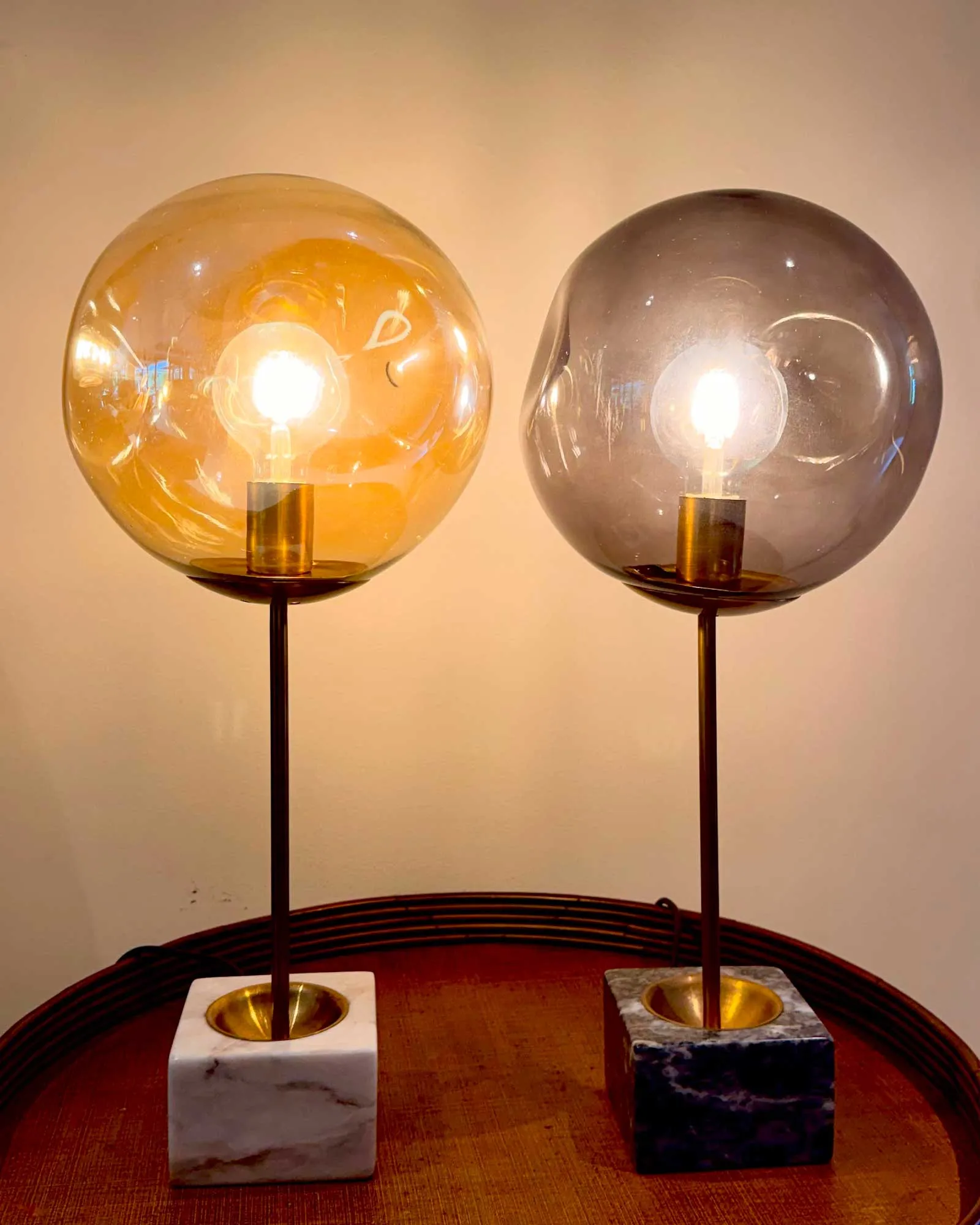 Two globe lamps with lit bulbs on tall metal stands, one with a white marble base and the other with a dark stone base on a wooden table.