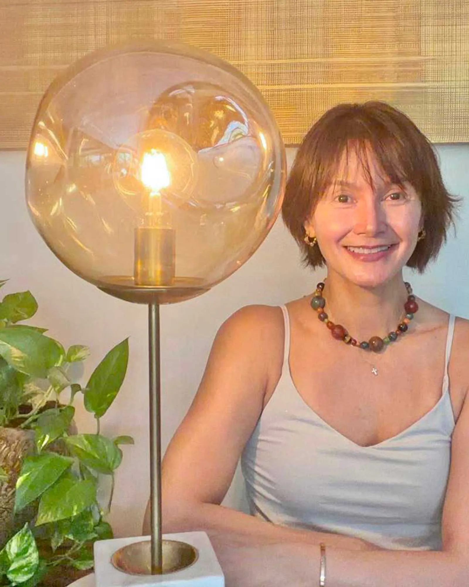 Smiling woman with short brown hair and a beaded necklace sits at a table next to a large glass globe lamp and leafy plants behind her.