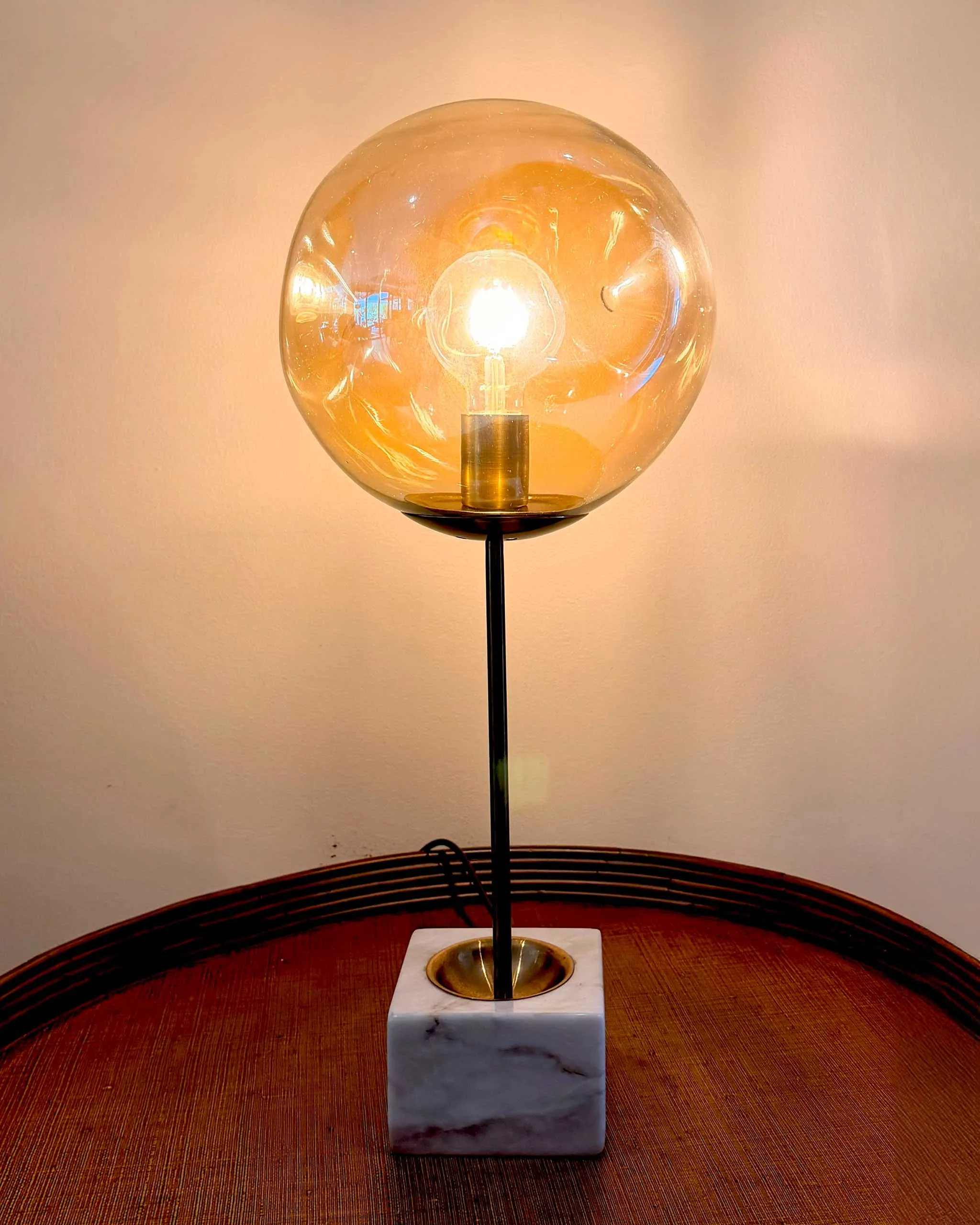 Table lamp with a clear glass globe surrounding a glowing bulb on a marble square base, on a wooden table against a beige wall.