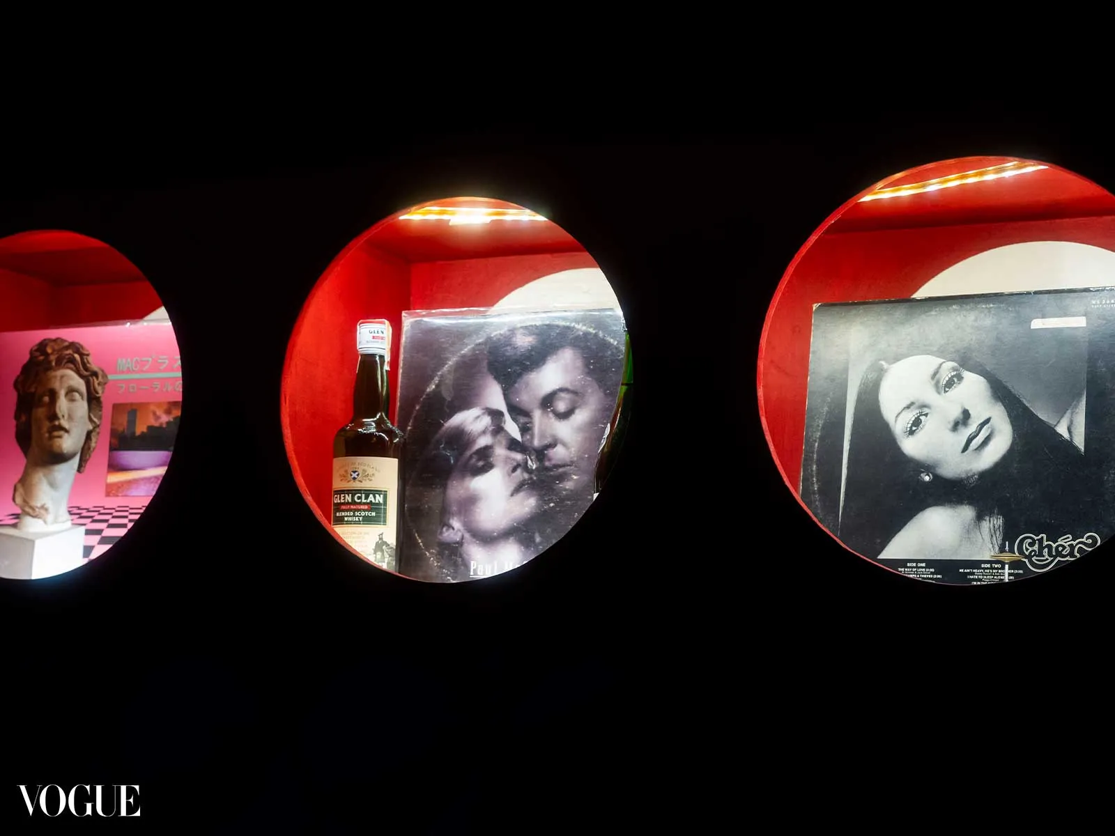 Three red circular display shelves on black, showing a statue with a pink poster, a whiskey bottle over a vintage photo, and a black-and-white portrait; Vogue logo.