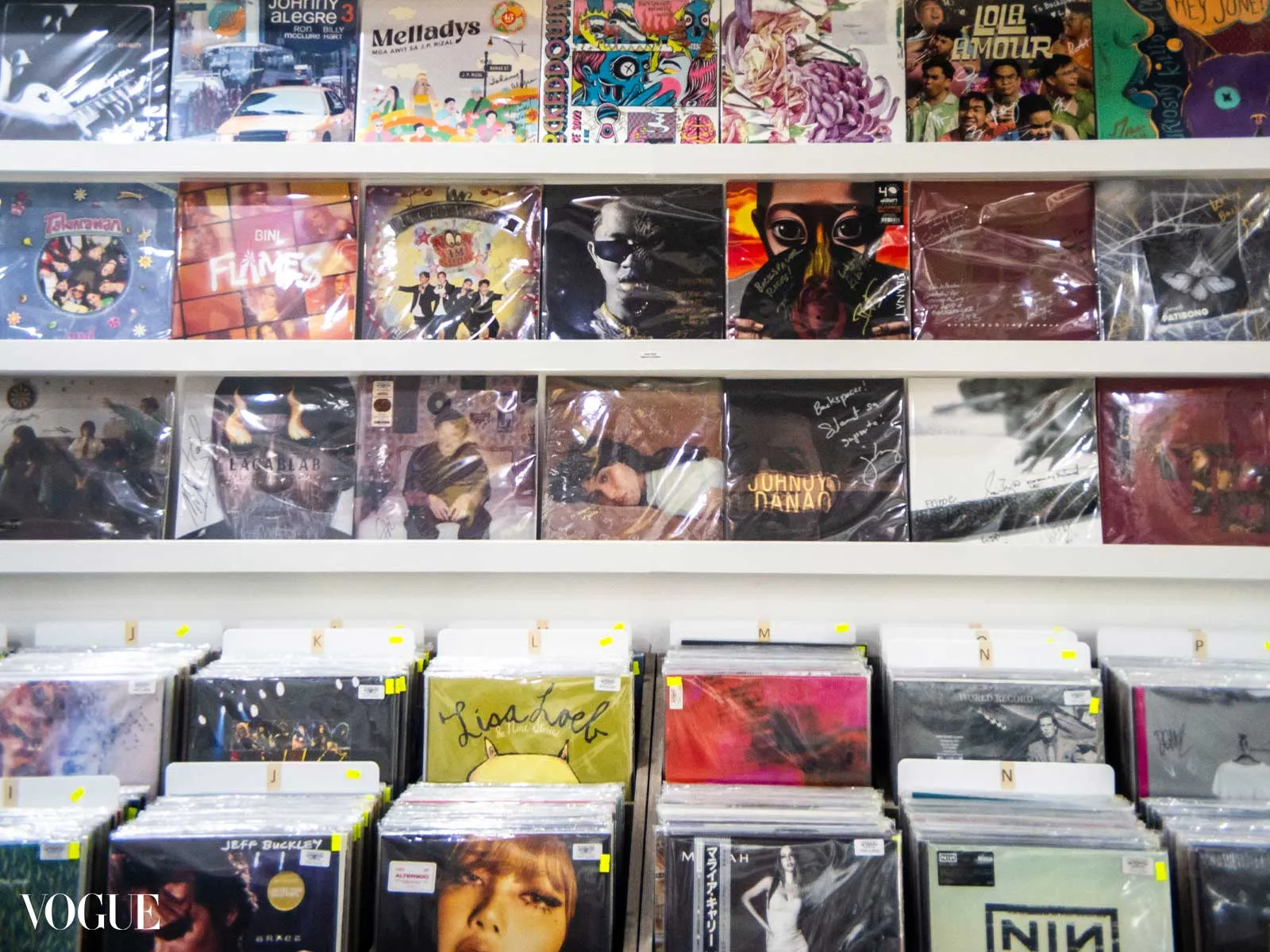 Rows of vinyl record sleeves on white shelves in a music store, colorful album art visible on top shelves and lower bins nearby