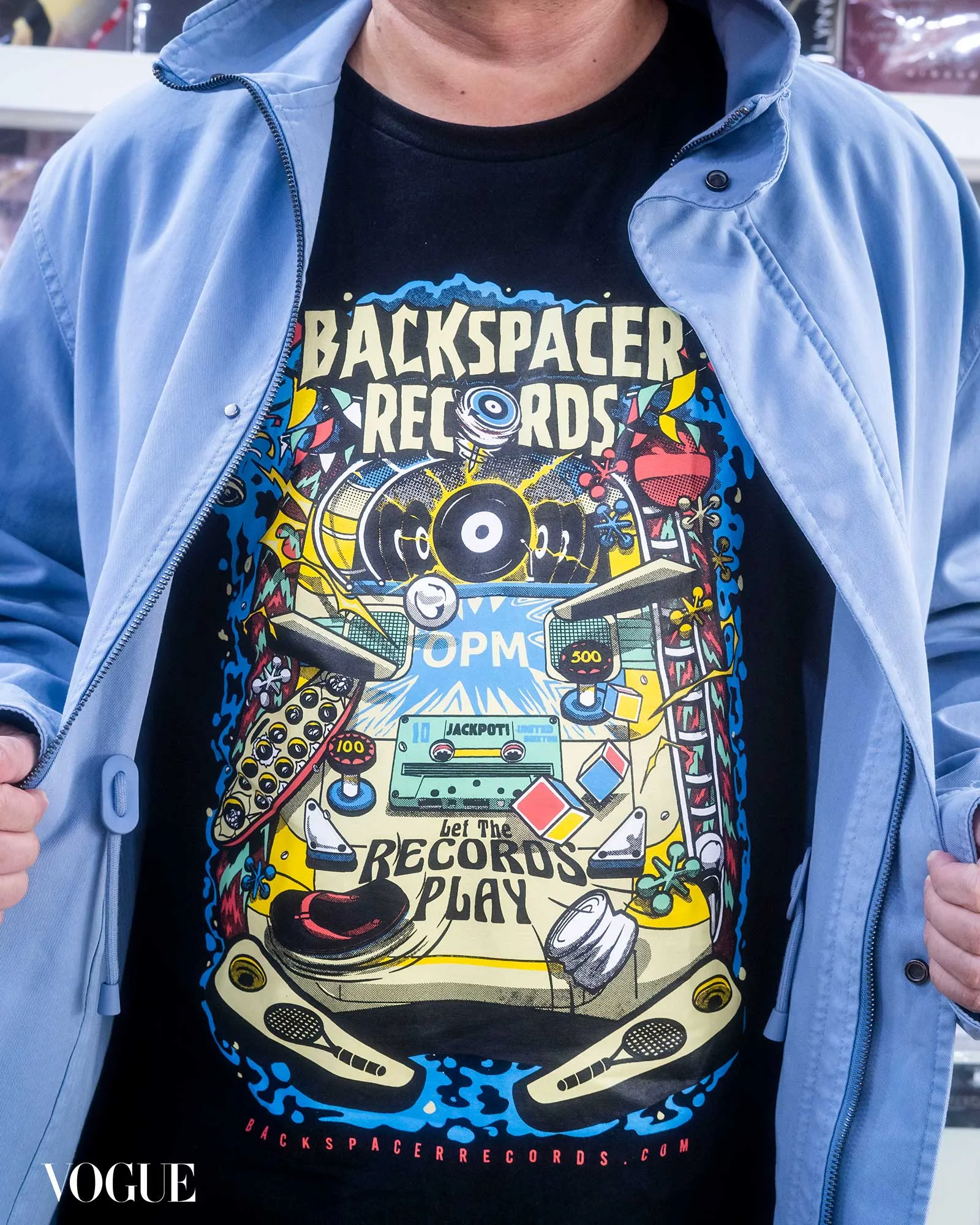 Person wearing a black graphic t‑shirt with a retro cassette design and 'BACKSPACER RECORDS' text, blue jacket over it, Vogue watermark bottom left.