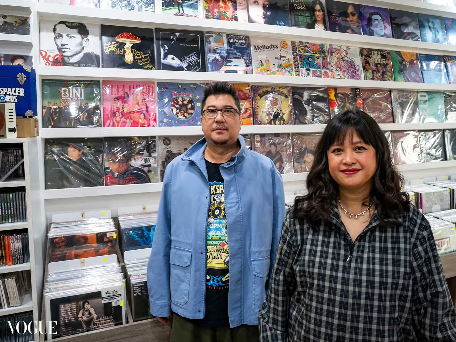 Two people pose in a vinyl record shop in front of wall shelves filled with colorful album covers behind them inside a music store.