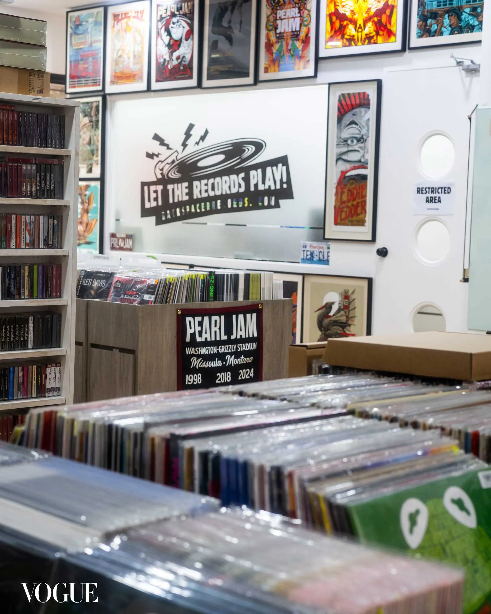 Interior of a record shop with stacked vinyl records in the foreground and framed posters on the walls; a large sign depicts a spinning record and reads 'Let the Records Play'