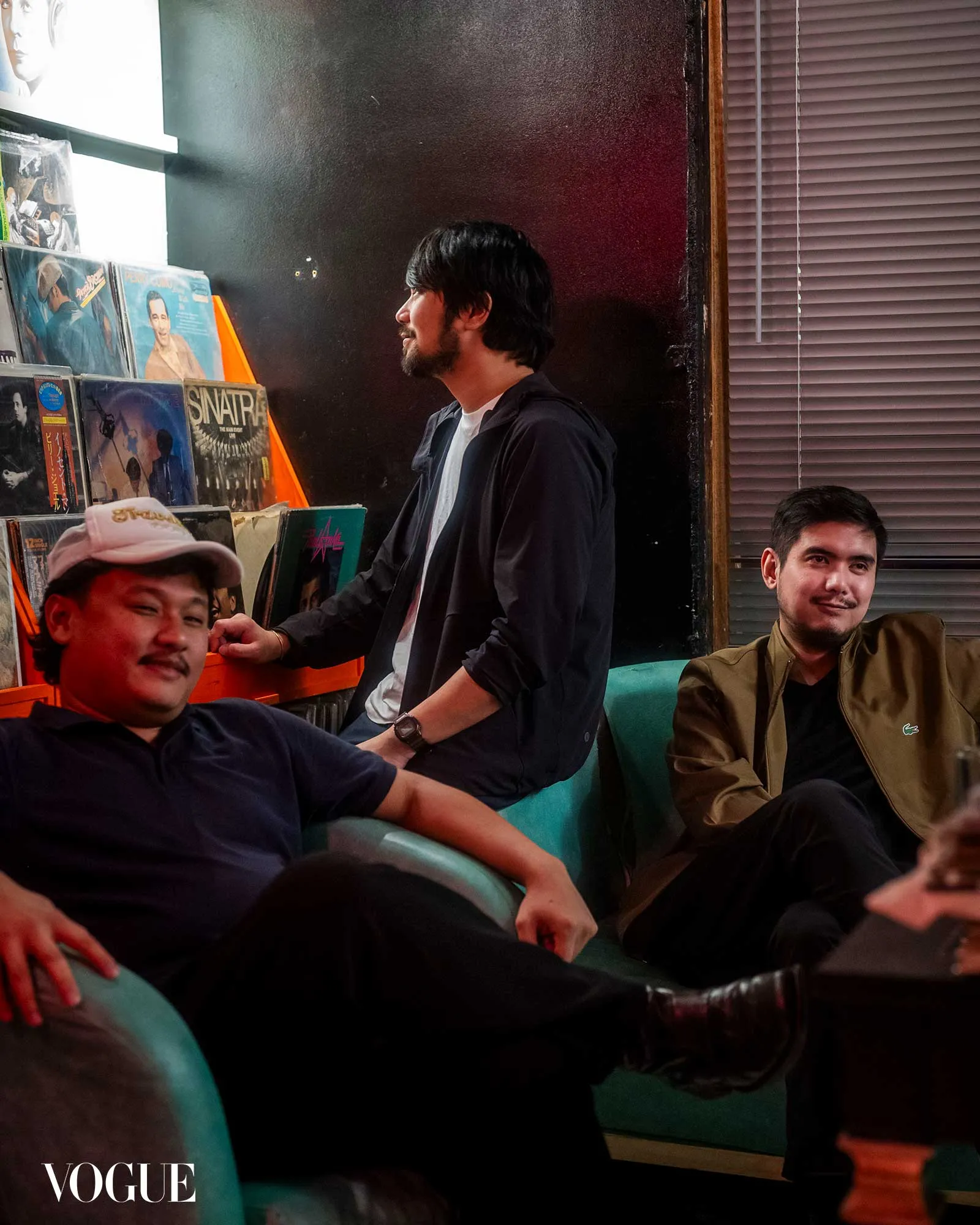 Man with a beard in a white shirt and dark jacket stands beside a vinyl record rack while two other men sit on a teal couch in a dim room.