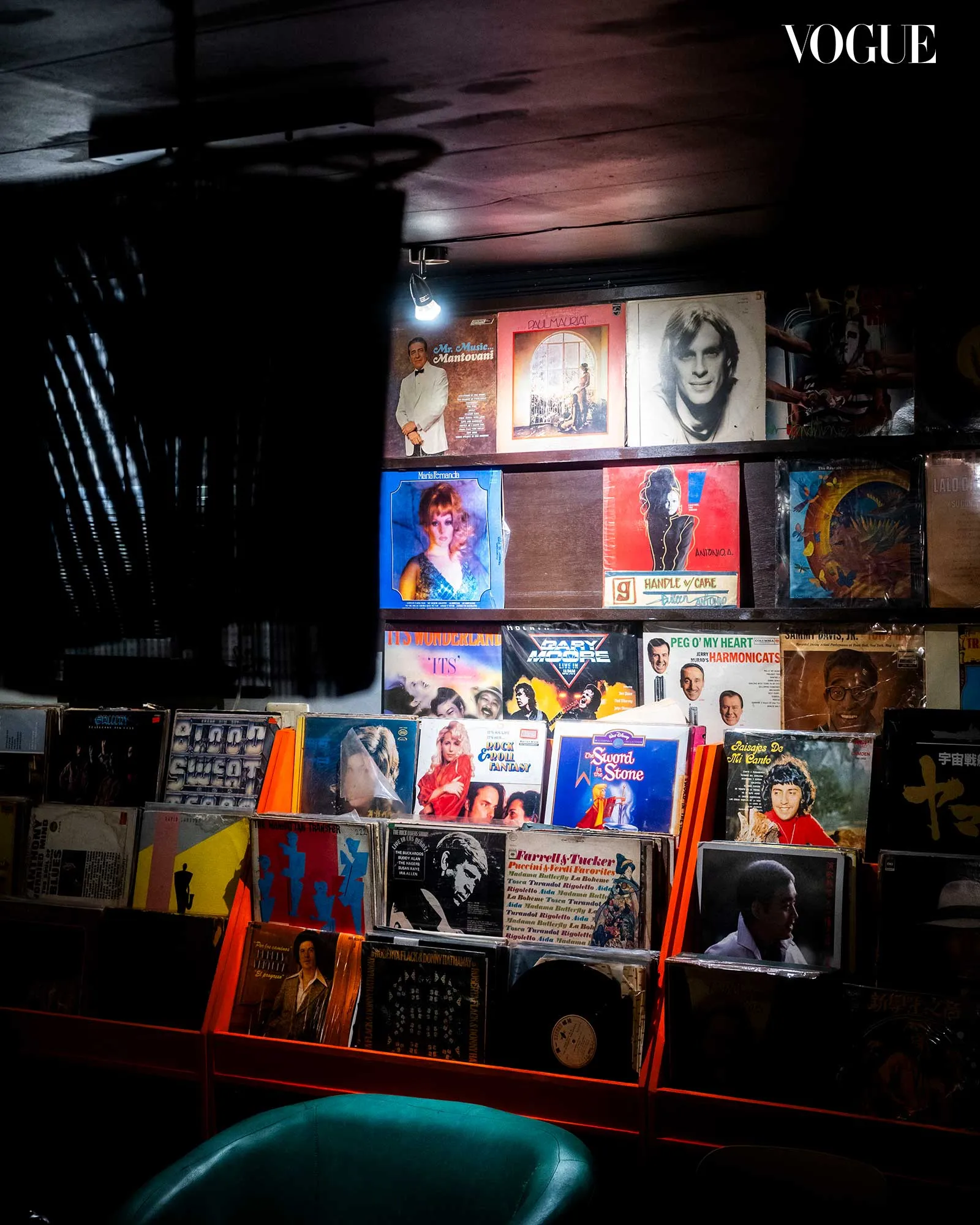 Dimly lit wall of vinyl records and vintage posters with faces, Vogue logo visible in the top-right corner; teal chair in foreground.