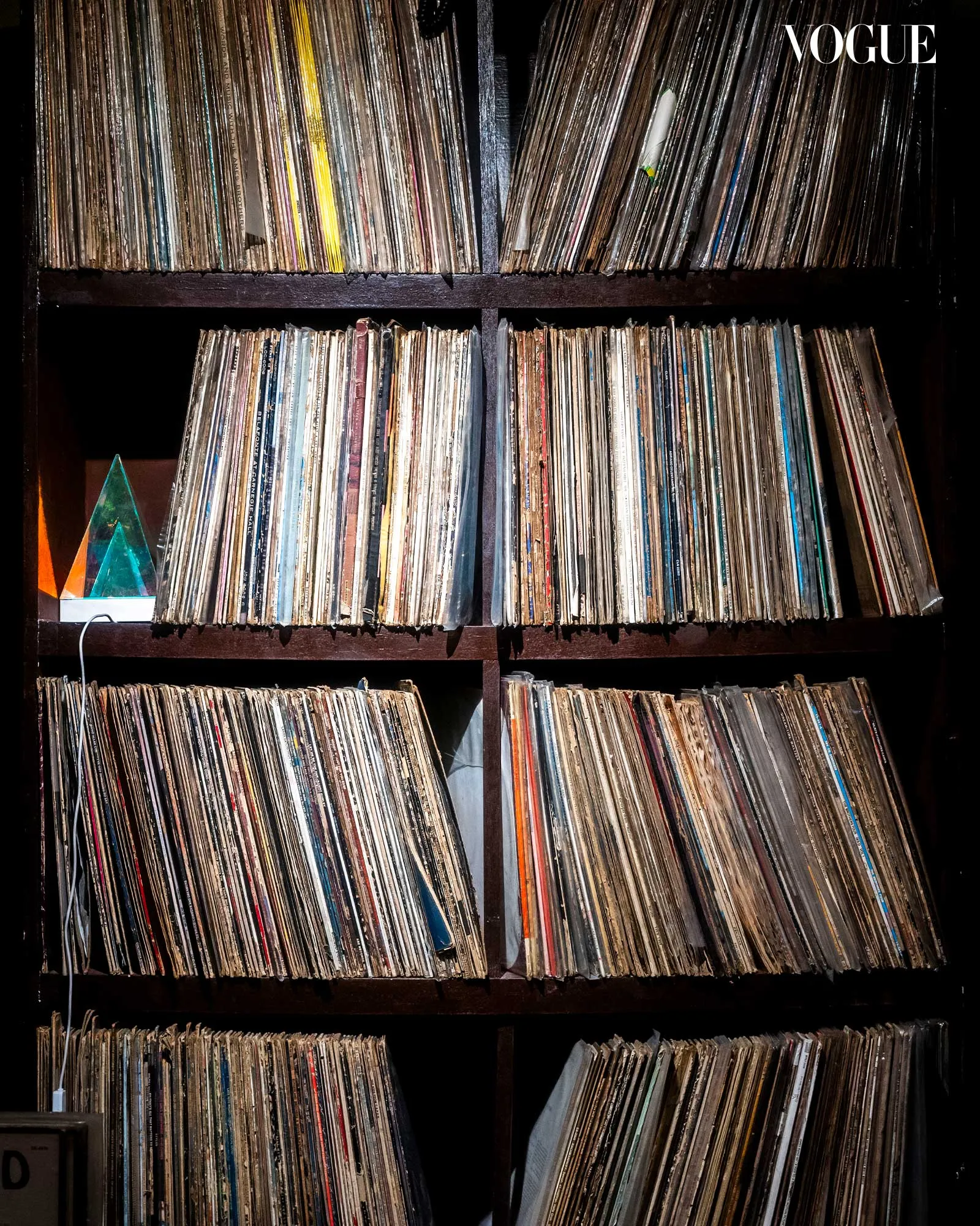 Rows of weathered vinyl record sleeves on dark shelves, a Vogue logo in the top-right corner, conveying a vinyl collection.