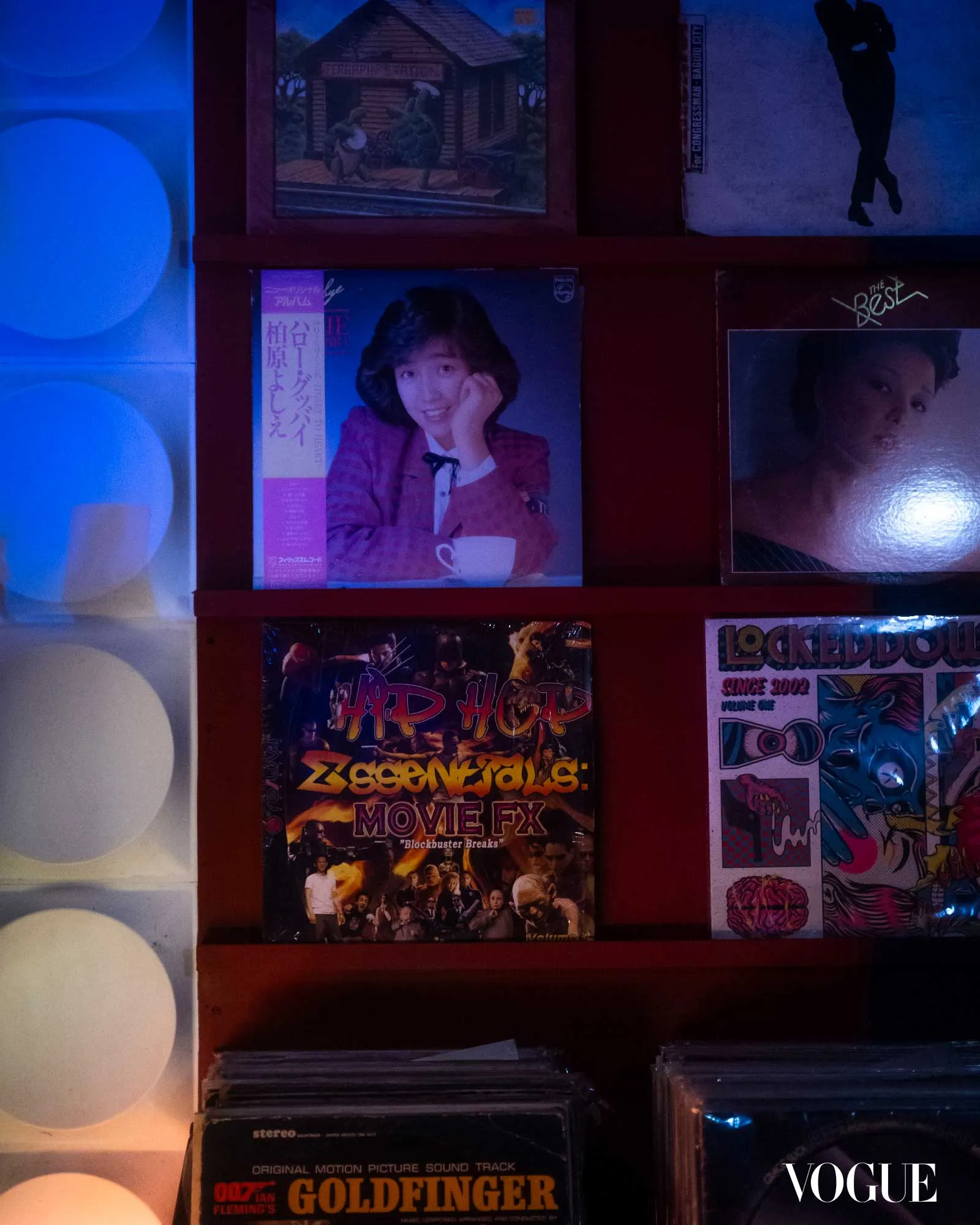 Wall-mounted display of vintage posters and album covers under blue lighting, including a James Bond Goldfinger poster centered lower row and Vogue logo at bottom right in a retro shop setting.