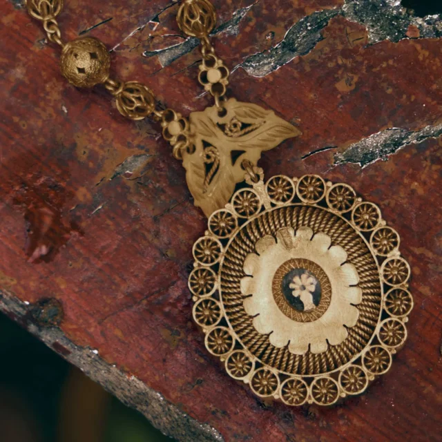 Intricate gold jewelry pendant with circular filigree design resting on a reddish stone surface, close-up view.