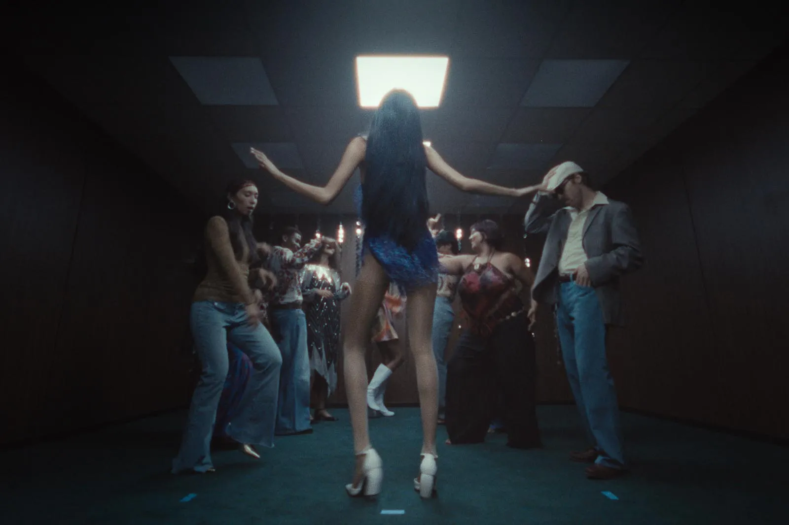 Woman in blue dress dancing with arms outstretched on a crowded dance floor, back to camera in a dim club setting.