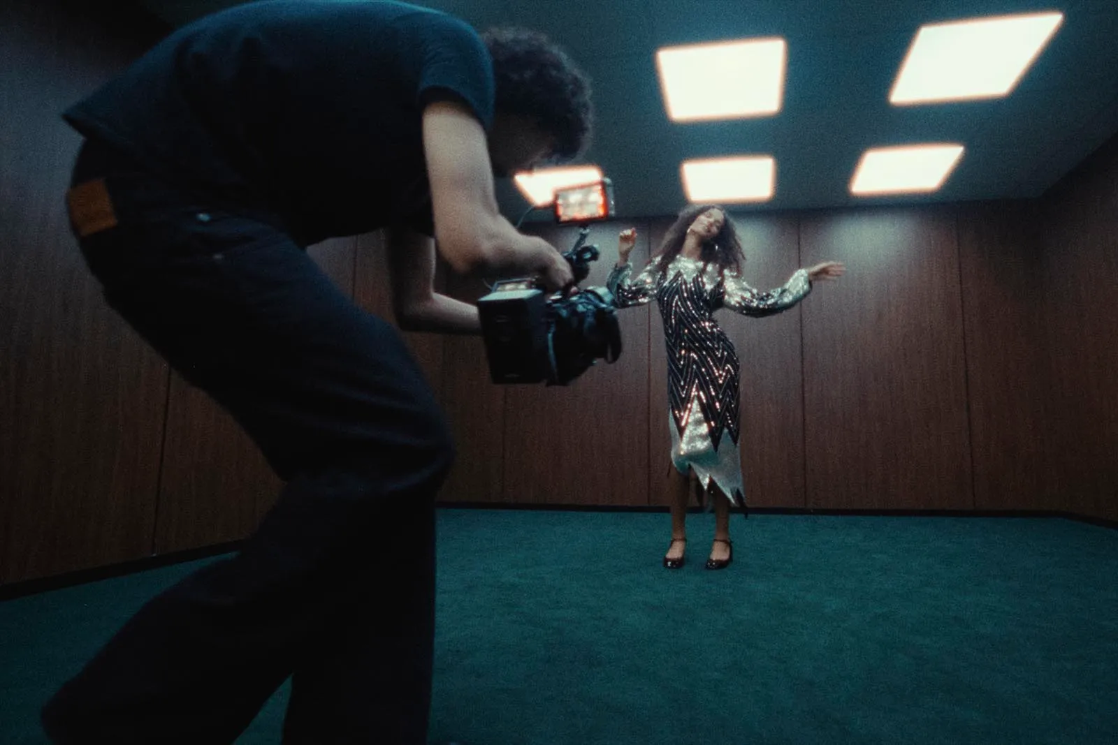 Photographer films a woman in a sequined zigzag dress in a dim, wood-paneled room with bright ceiling lights.
