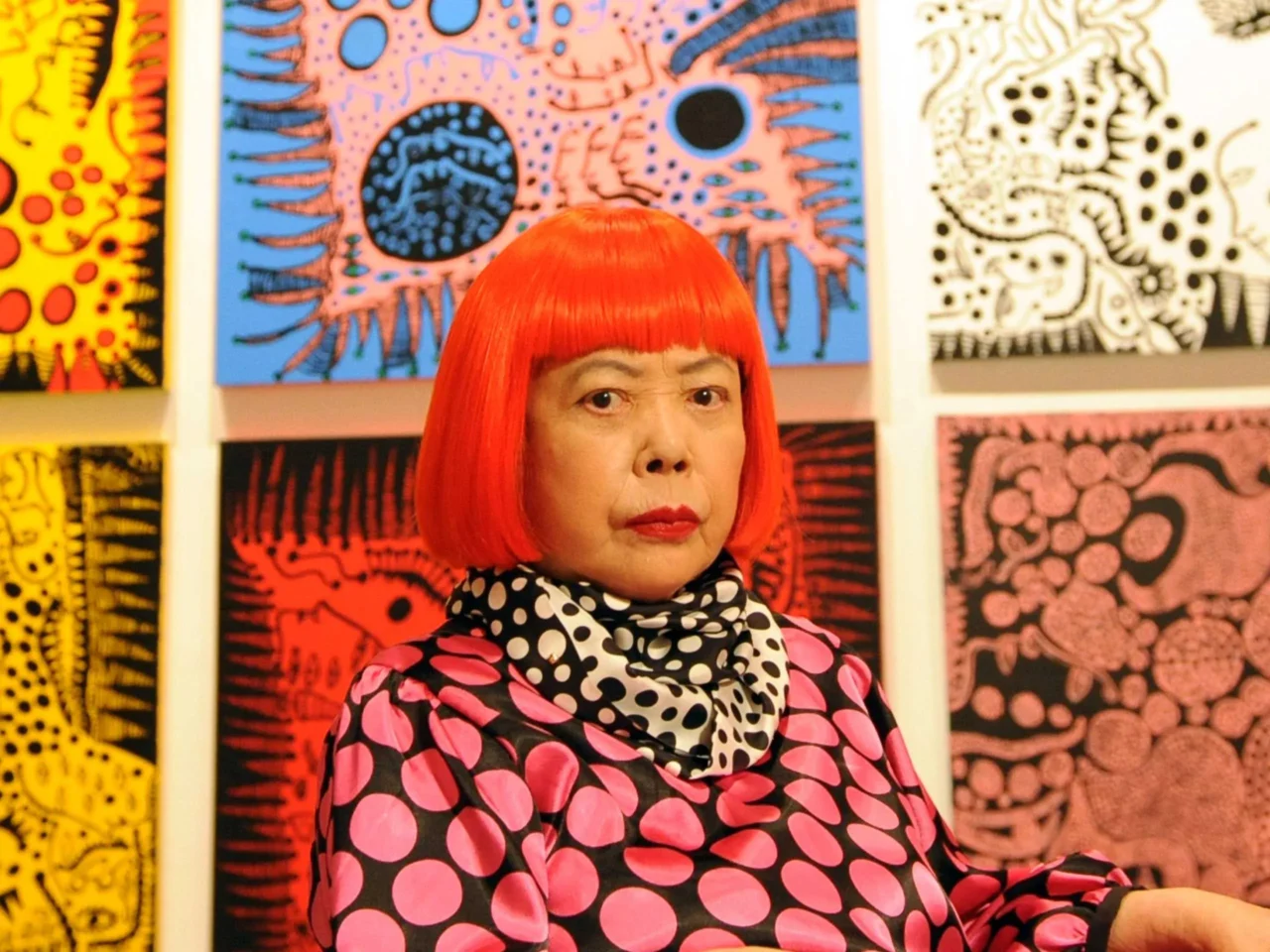 Portrait of a person with a bright orange bob haircut wearing a pink and black polka-dot outfit and a white-and-black polka scarf, standing in front of colorful abstract artwork.