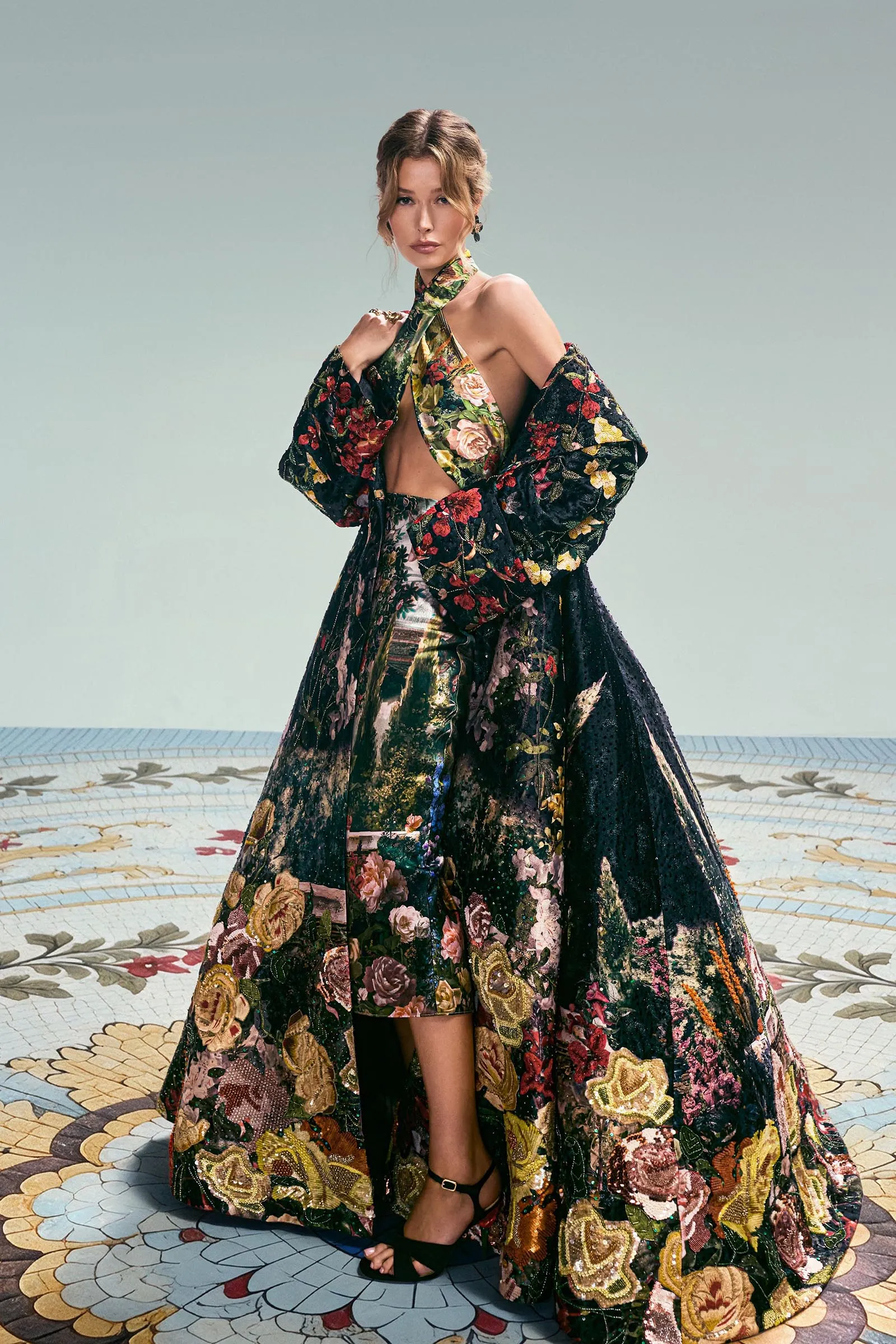 Woman in a dramatic, floor-length floral gown with a matching cape, posing gracefully on a patterned mosaic floor.