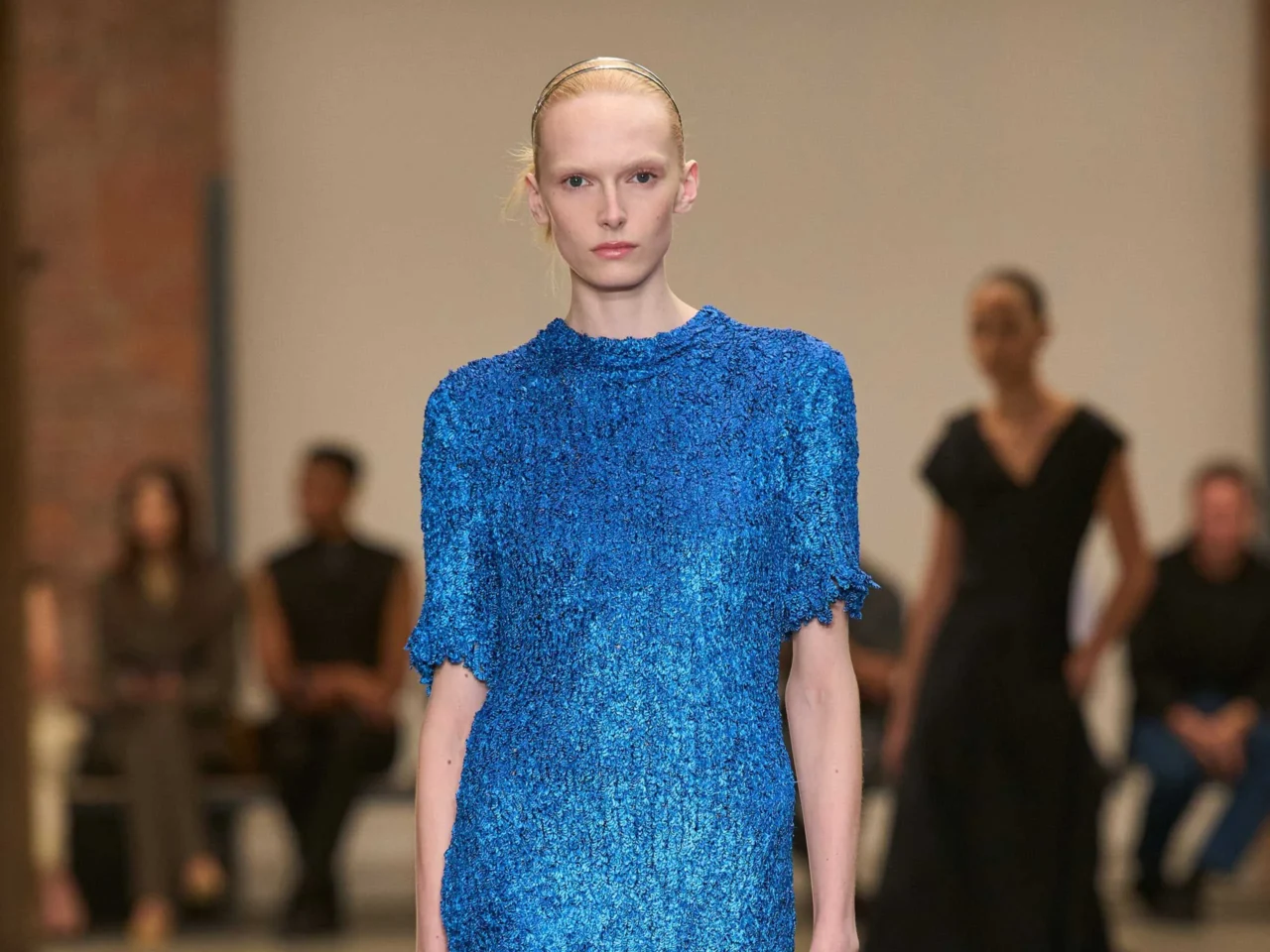 Model walks the runway in a bright blue textured dress; blurred audience in the background,