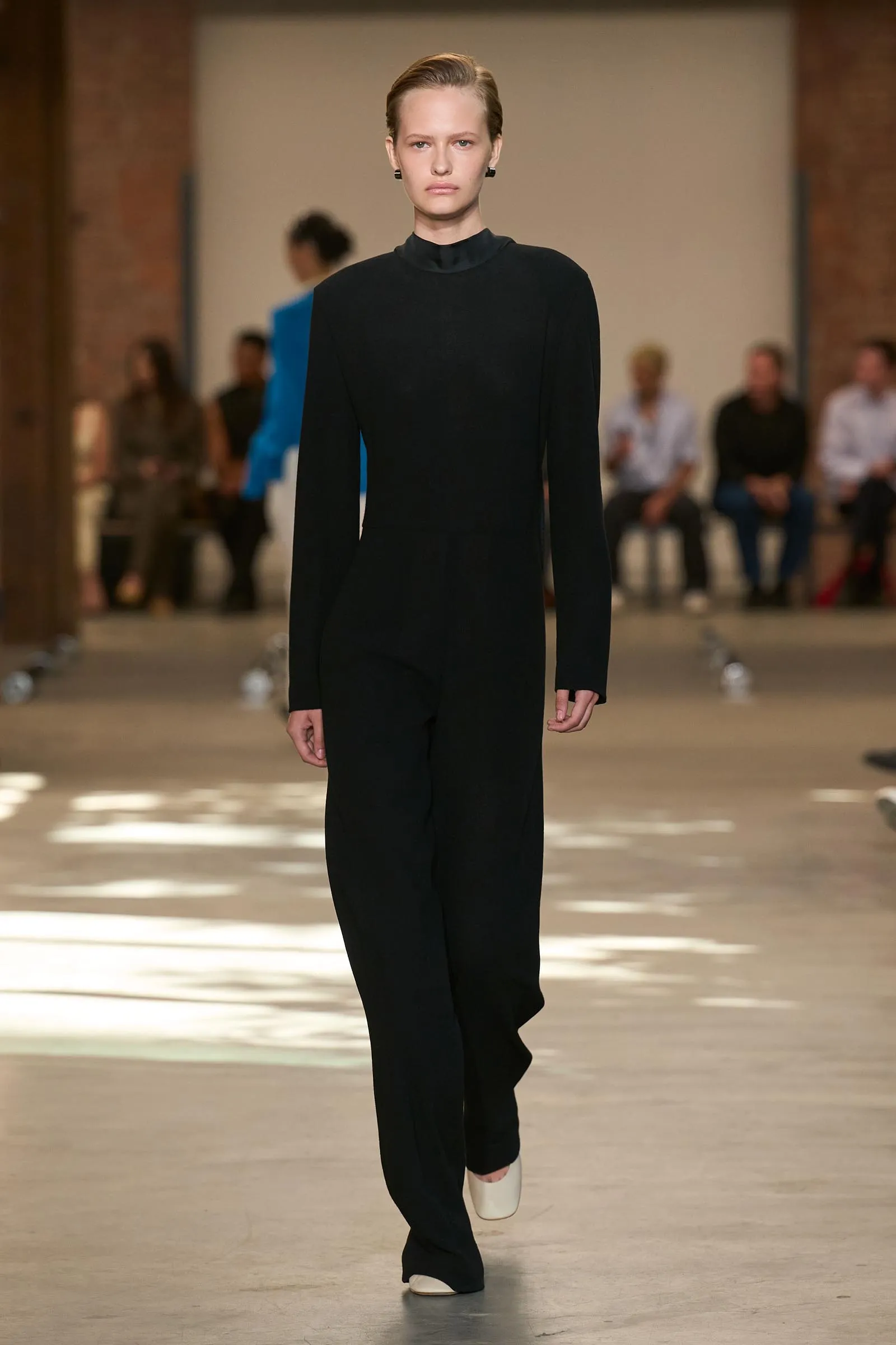 Model walks the runway in a bright blue textured dress; blurred audience in the background,