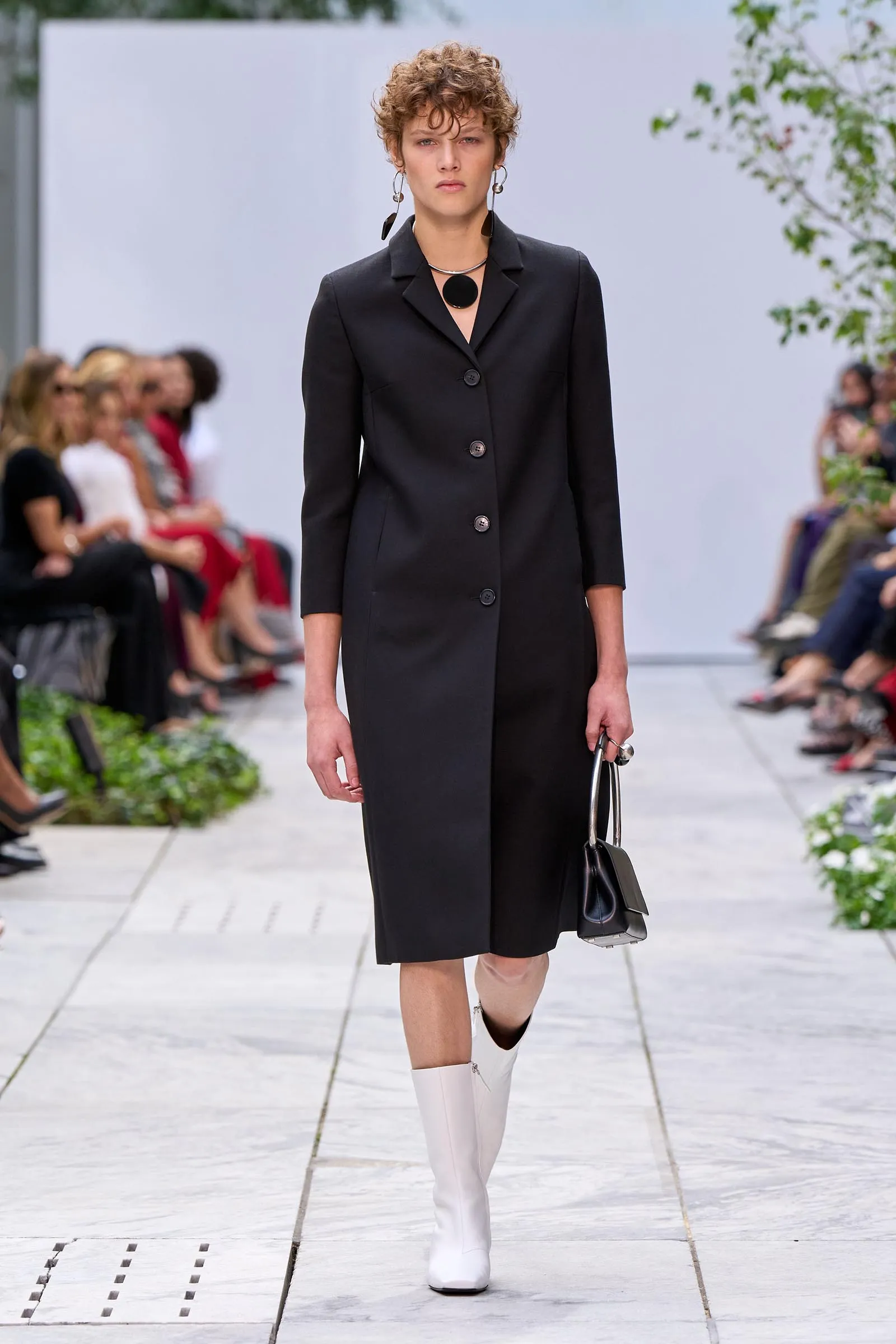 Model walking a runway in a black sleeveless V-neck dress with a silver choker and bold red lipstick, blurred spectators in the background.
