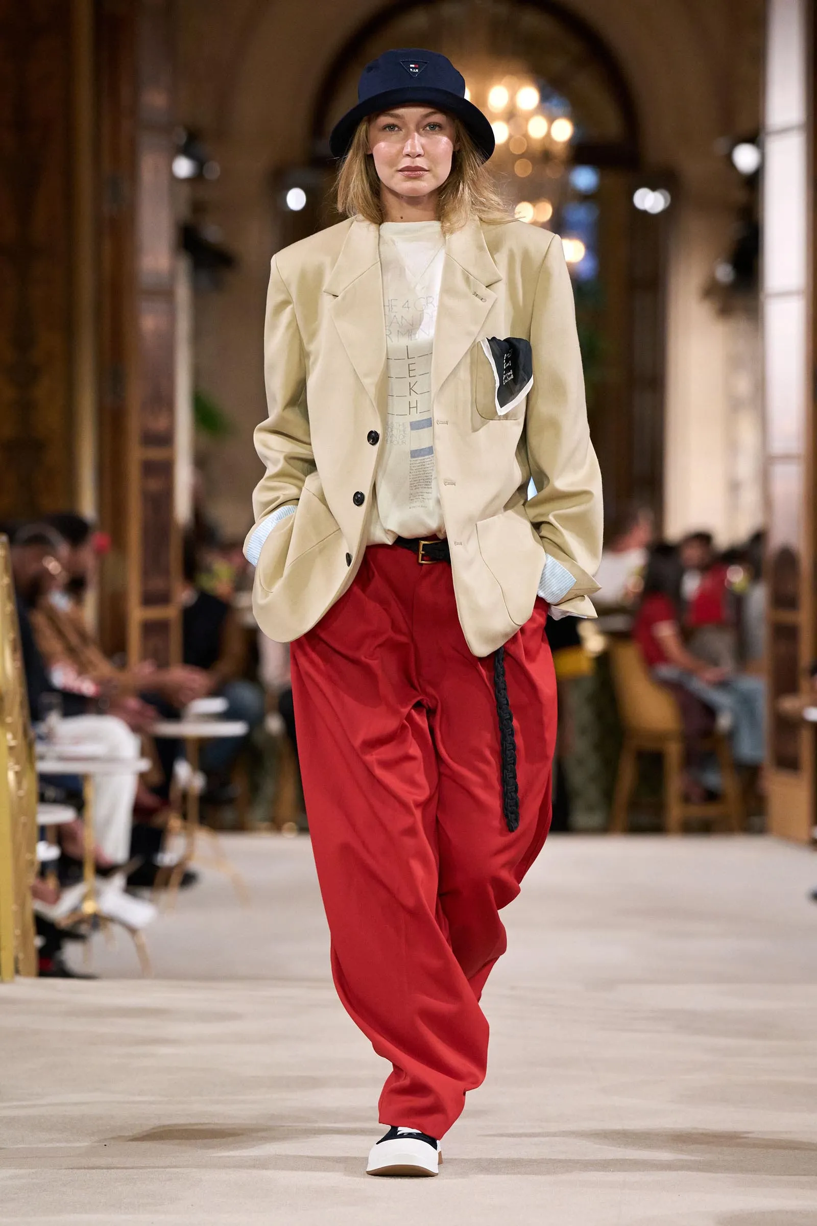 Female model walking a fashion runway in a beige blazer, white graphic tee, and bright red baggy pants, wearing a dark bucket hat. Background shows an indoor venue with seated audience and warm lighting.