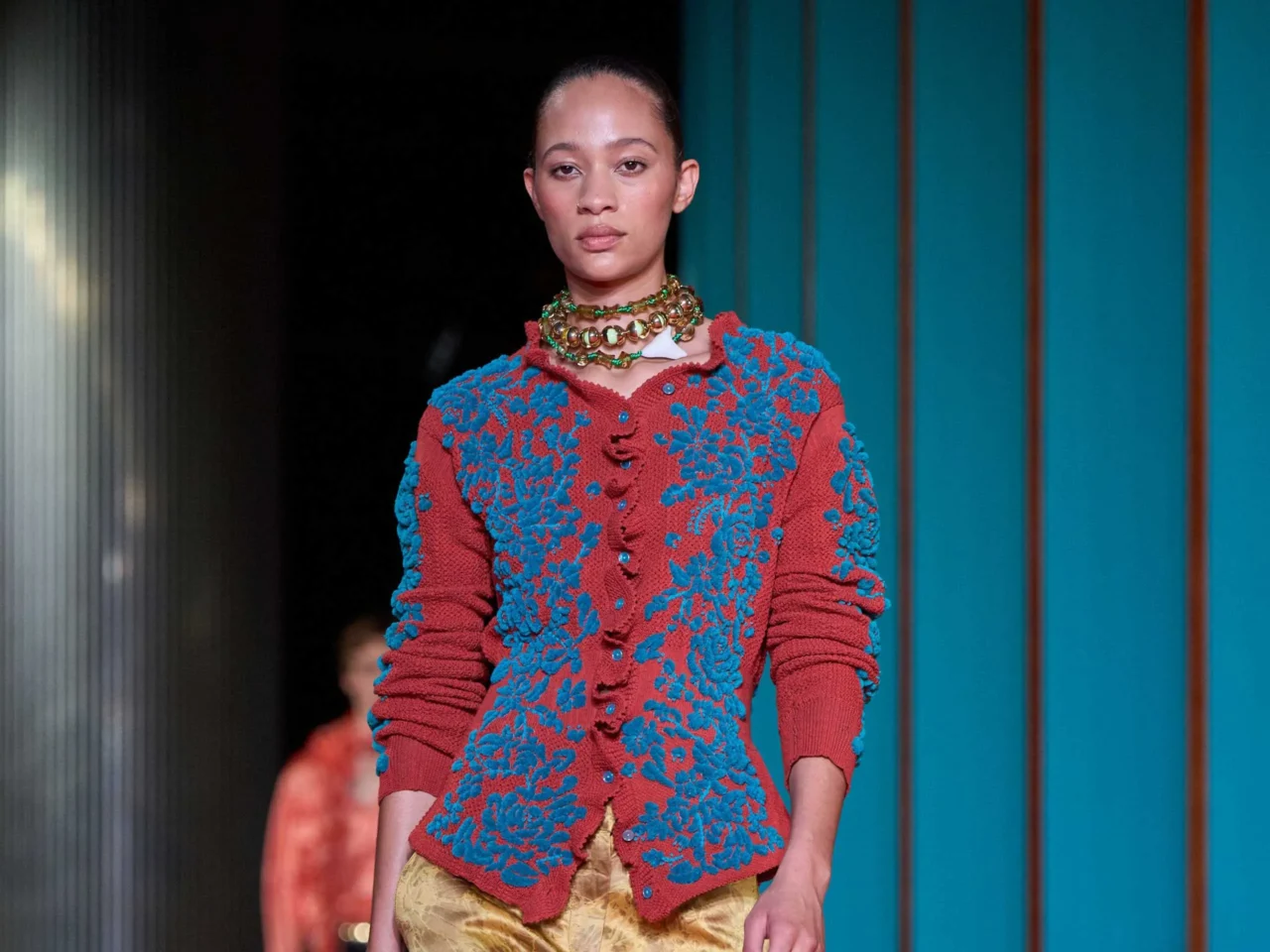 Portrait of a woman with dark hair wearing a green knit sweater with purple floral embroidery and a chunky beaded necklace, against teal panel backdrop.