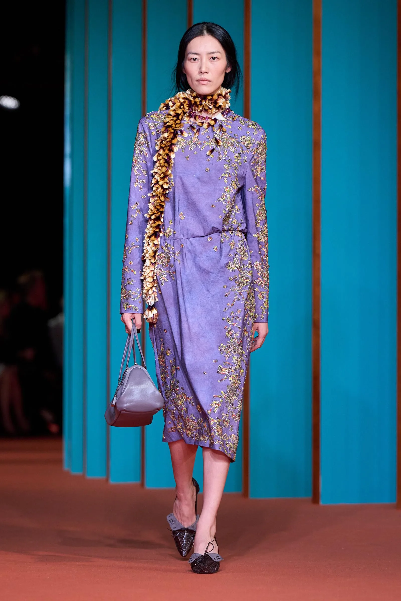 Portrait of a woman with dark hair wearing a green knit sweater with purple floral embroidery and a chunky beaded necklace, against teal panel backdrop.