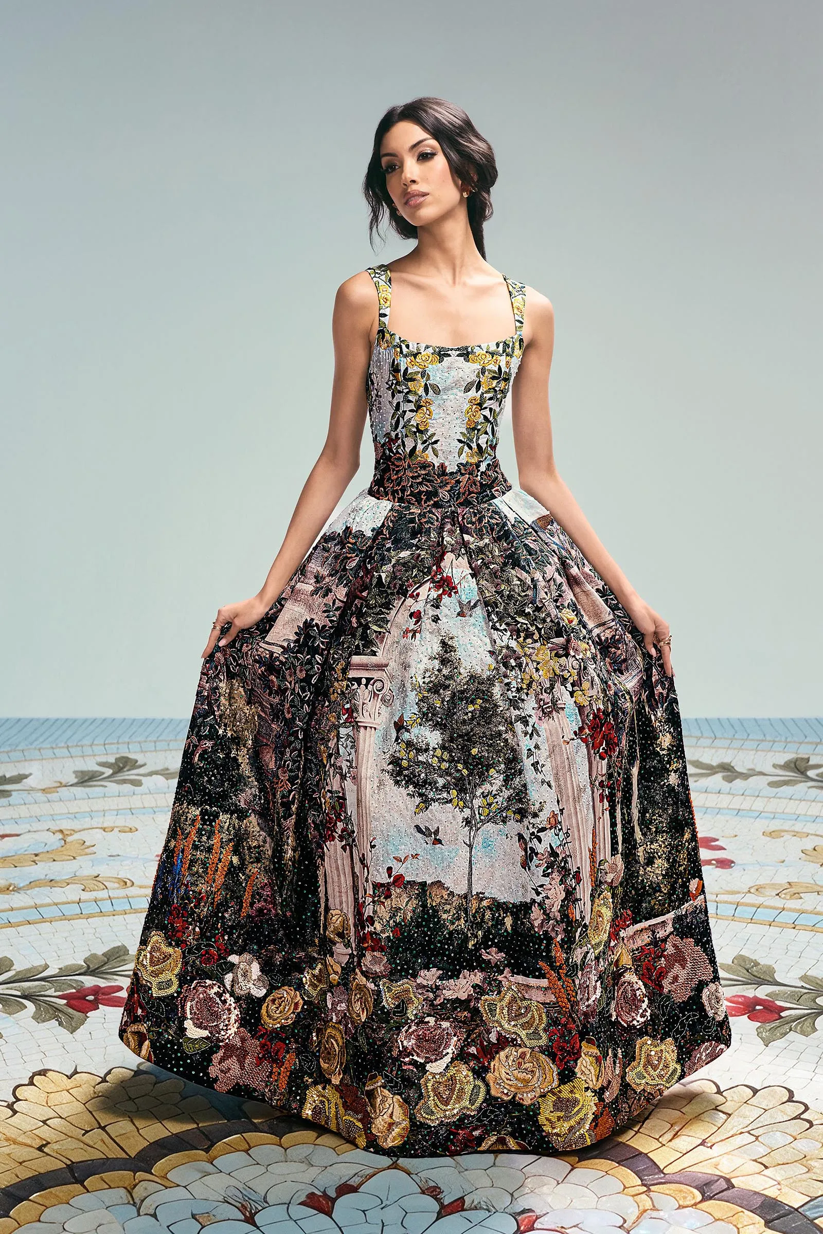 Model wearing a full-length floral-embroidered gown, standing with hands at her sides against a pale backdrop.
